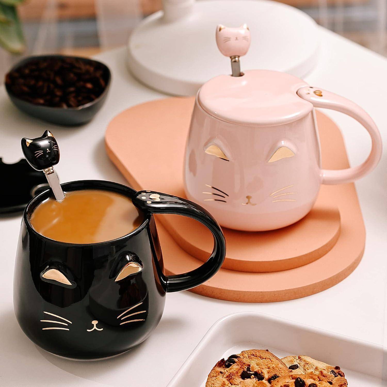 Zwei Katzen-Tassen, eine schwarz und eine rosa, mit passenden Löffeln, gefüllt mit Kaffee. Sie stehen auf einem Tisch neben Keksen.