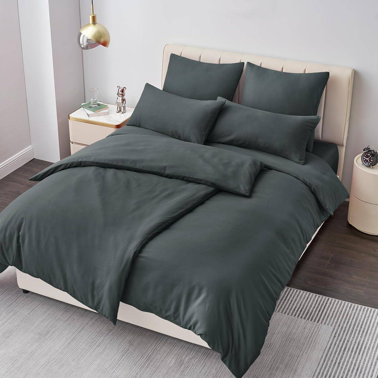Ein ordentlich gemachtes Bett mit dunkelgrauer Bettwäsche, einschließlich Kissen und Bettdecke, ist vor einem hellgefärbten Kopfteil platziert. Auf dem Nachttisch stehen eine Lampe und kleine Dekorationsgegenstände.