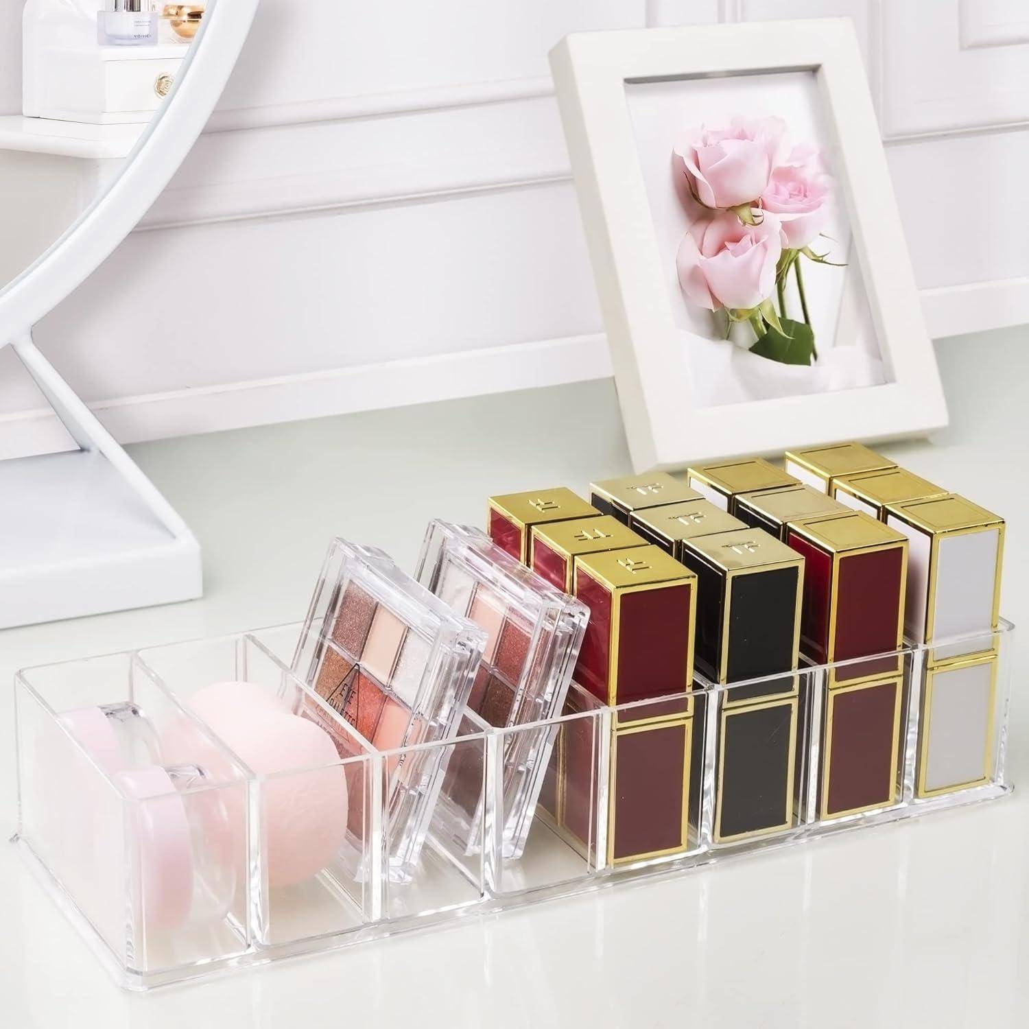 Ein durchsichtiger Organizer enthält verschiedene Lippenstifte, Rouge und einen Make-up-Schwamm auf einer Schminkommode. Ein gerahmtes Foto mit rosa Rosen steht dahinter.