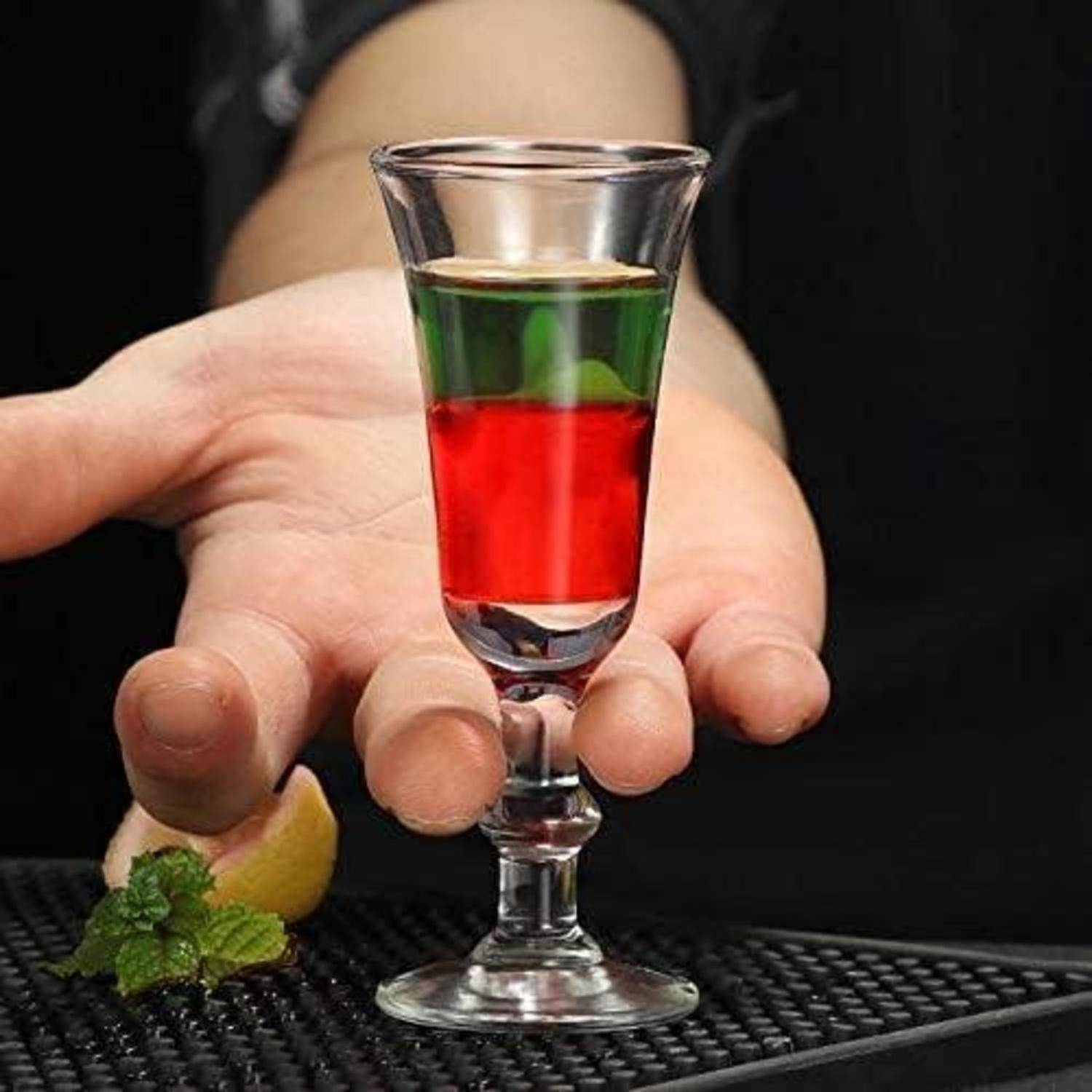 Eine Person hält einen geschichteten Cocktail in einem kleinen Glas mit roten und grünen Flüssigkeiten. Eine Zitronenscheibe und Minze sind auf der Oberfläche darunter sichtbar.