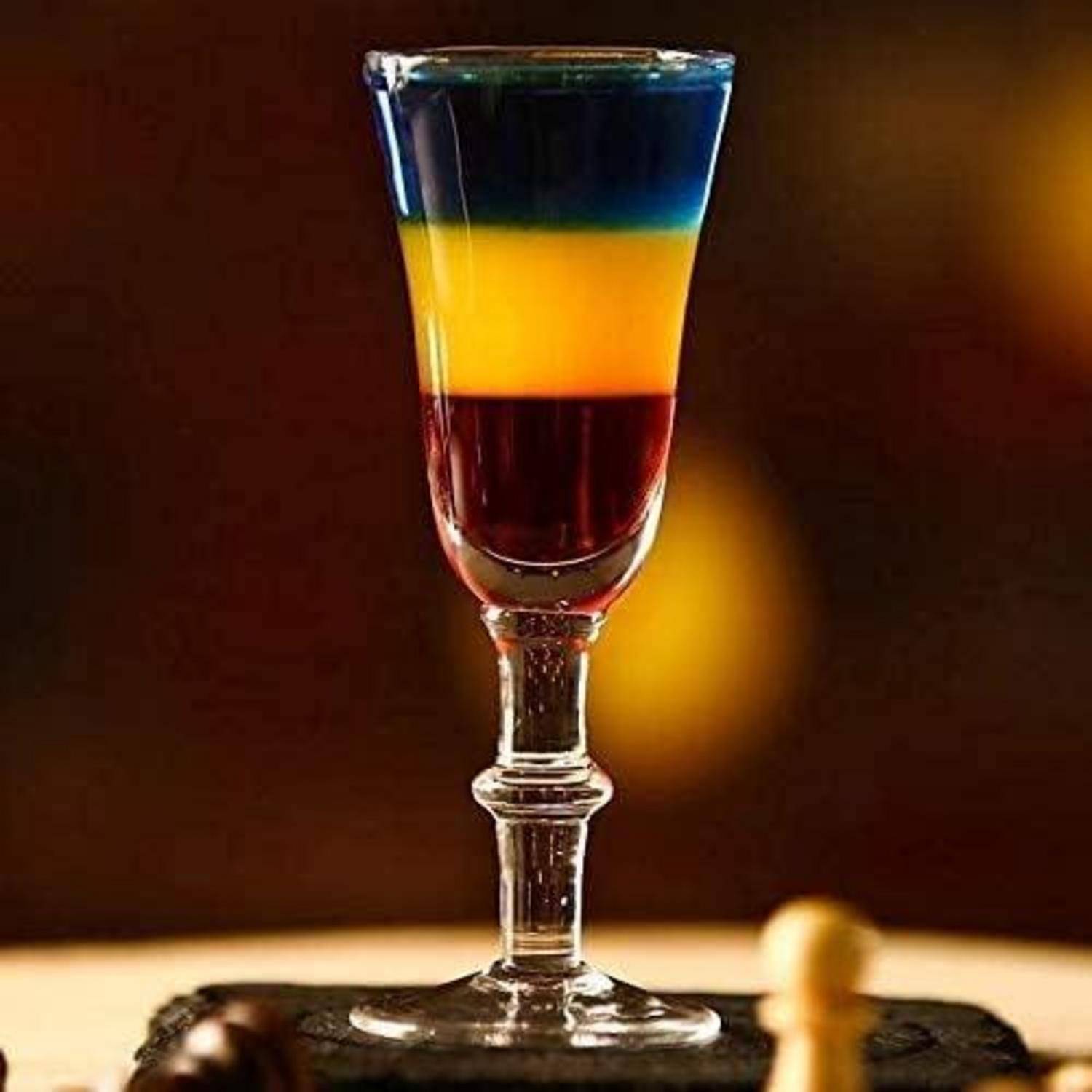 Ein geschichteter Cocktail in einem kleinen Glas zeigt deutliche blaue, gelbe und rote Schichten, vor einem unscharfen Hintergrund.
