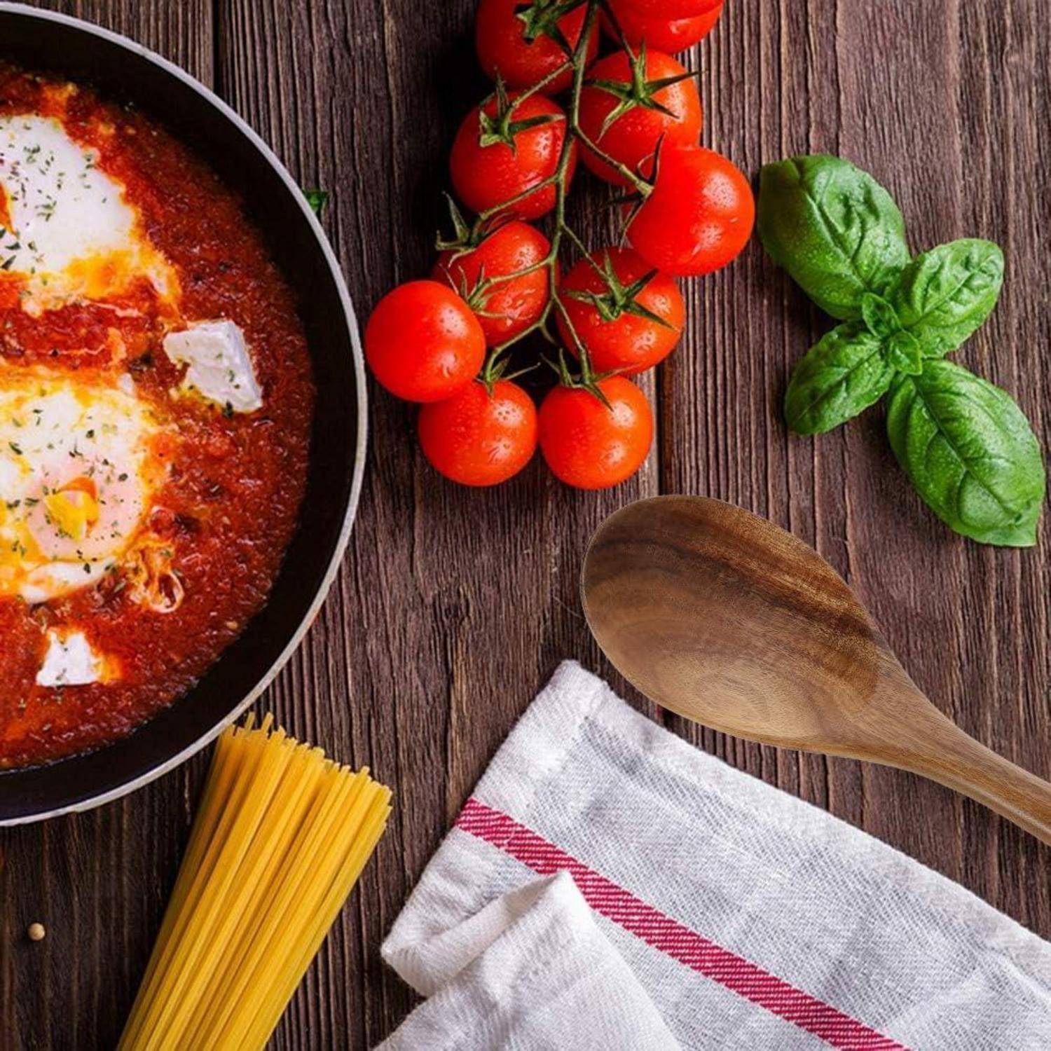 Eine Pfanne mit Tomatensoße, in der Eier garen, steht auf einem Holztisch. Daneben liegen frische Kirschtomaten, Basilikumblätter, ein Holzlöffel und rohe Spaghetti.