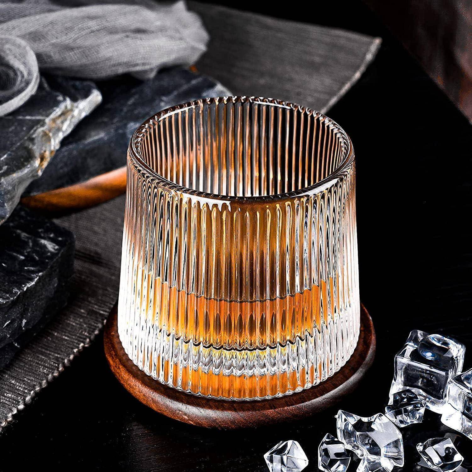 Ein Glas mit vertikalen Rillen, gefüllt mit einer braunen Flüssigkeit, steht auf einem Holzuntersetzer auf einer dunklen Oberfläche, umgeben von Eiswürfeln und Steinplatten.