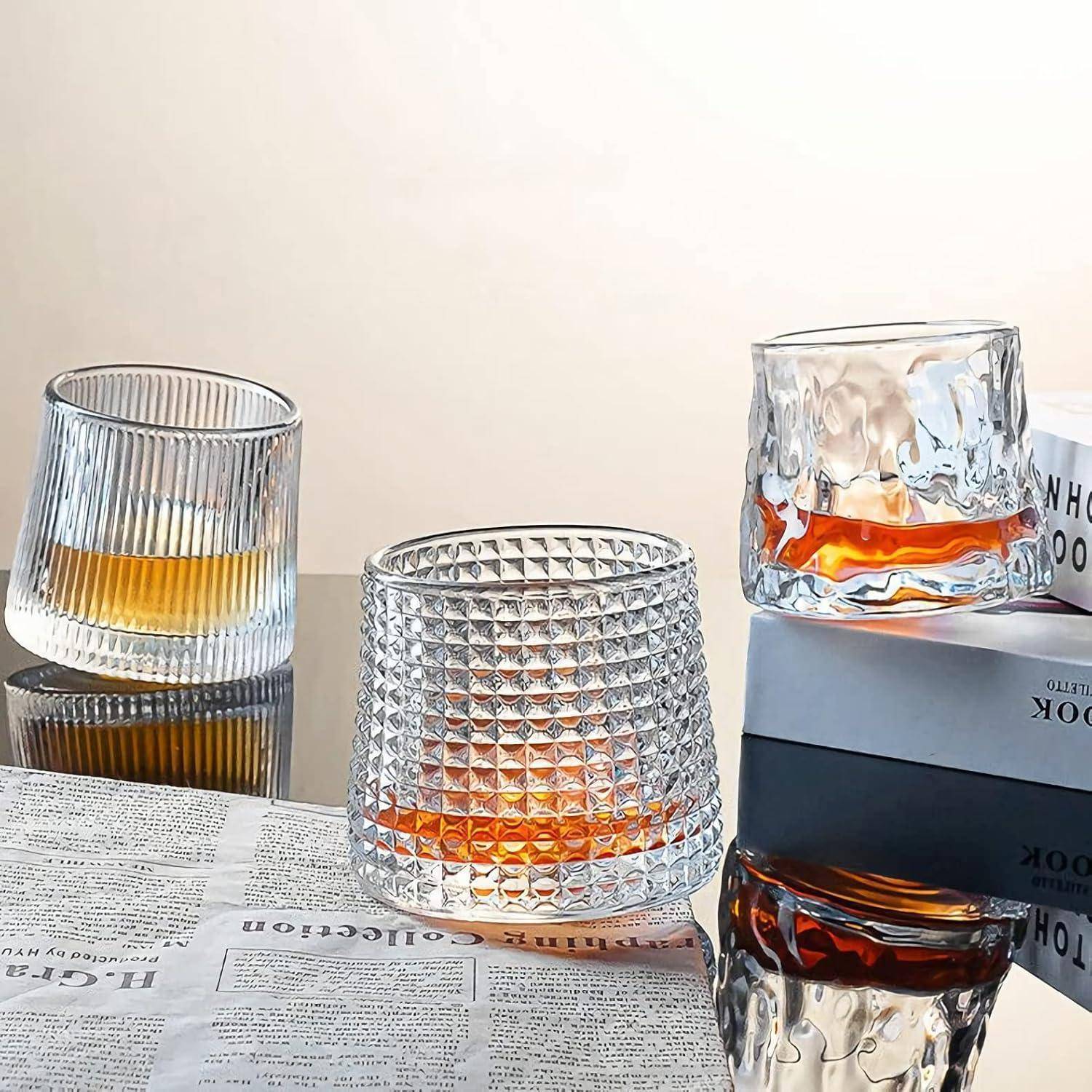 Drei strukturierte Whiskygläser mit unterschiedlichen Formen, die auf einer reflektierenden Oberfläche neben geöffneten Büchern und Papieren platziert sind.