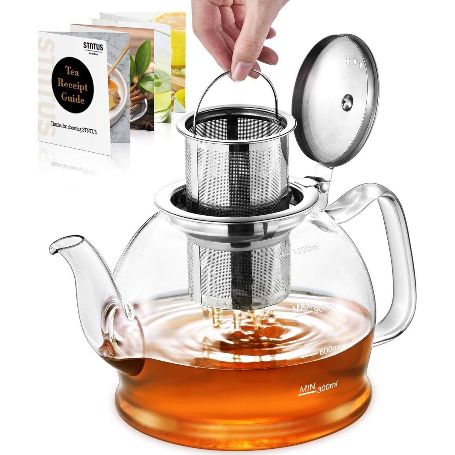 Eine Hand hebt einen Metallinfuser aus einer durchsichtigen Glaskanne, die bernsteinfarbenen Tee enthält. Die Teekanne hat Messskalen. Ein Heft mit dem Titel 