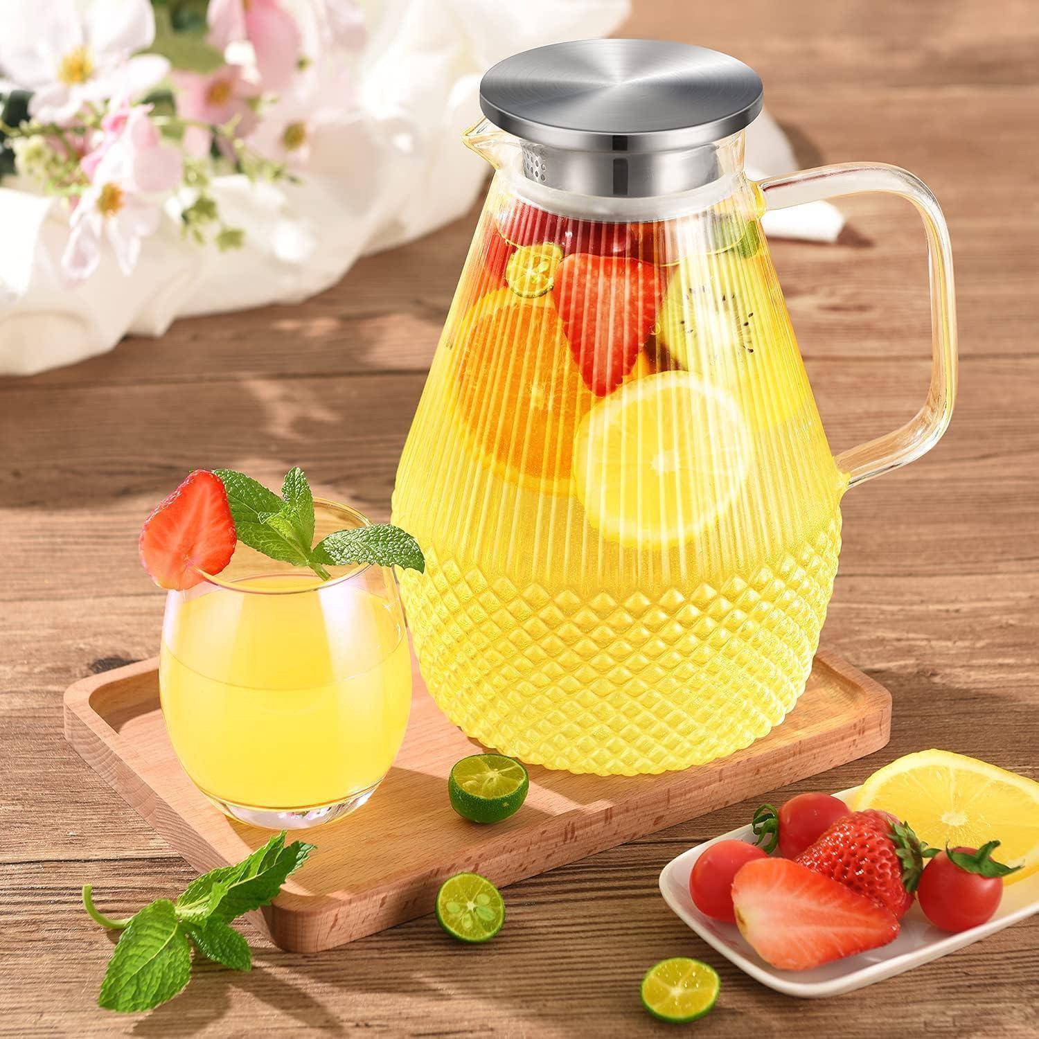 Eine klare Glaskanne, gefüllt mit zitrischen und beereninfundierten gelben Getränk, steht auf einem Holztablett mit einem passenden Glas und frischer Obstgarnitur.