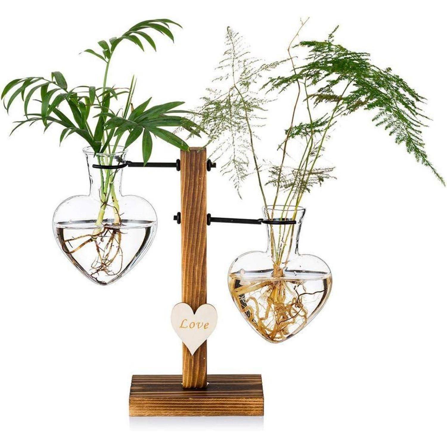 Herzförmige Glasvasen mit grünen Pflanzen sind auf einem Holzständer montiert. Ein kleines Holzherz an der Basis trägt das Wort 'Love'.