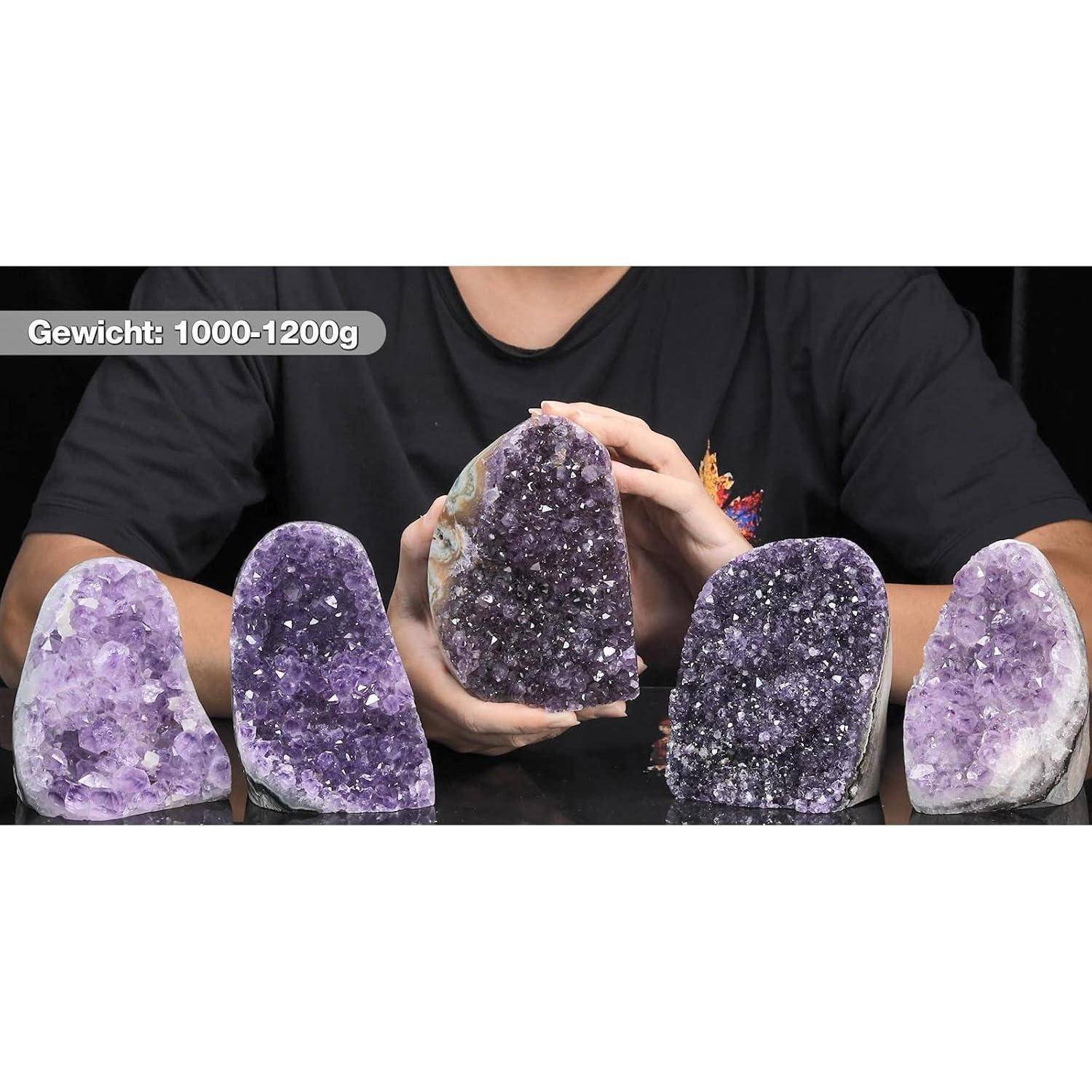 Eine Person, die eine große Amethyst-Geode umgeben von kleineren Amethyst-Geoden auf einer reflektierenden Oberfläche hält. Text lautet: 'Gewicht: 1000-1200g'.