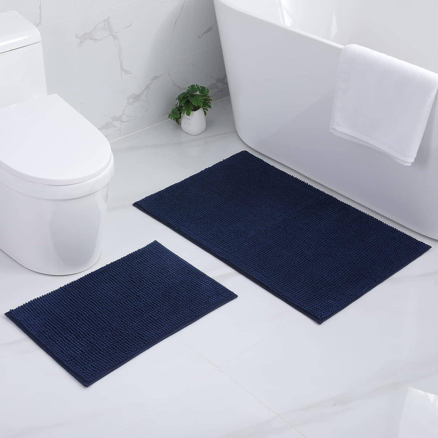 Eine Badezimmerszene mit einer weißen Toilette, einer kleinen Pflanze auf dem Boden, zwei marineblau-farbenen Badematte und einer Badewanne mit einem weiß-farbenen Handtuch, das über den Rand gehängt ist.