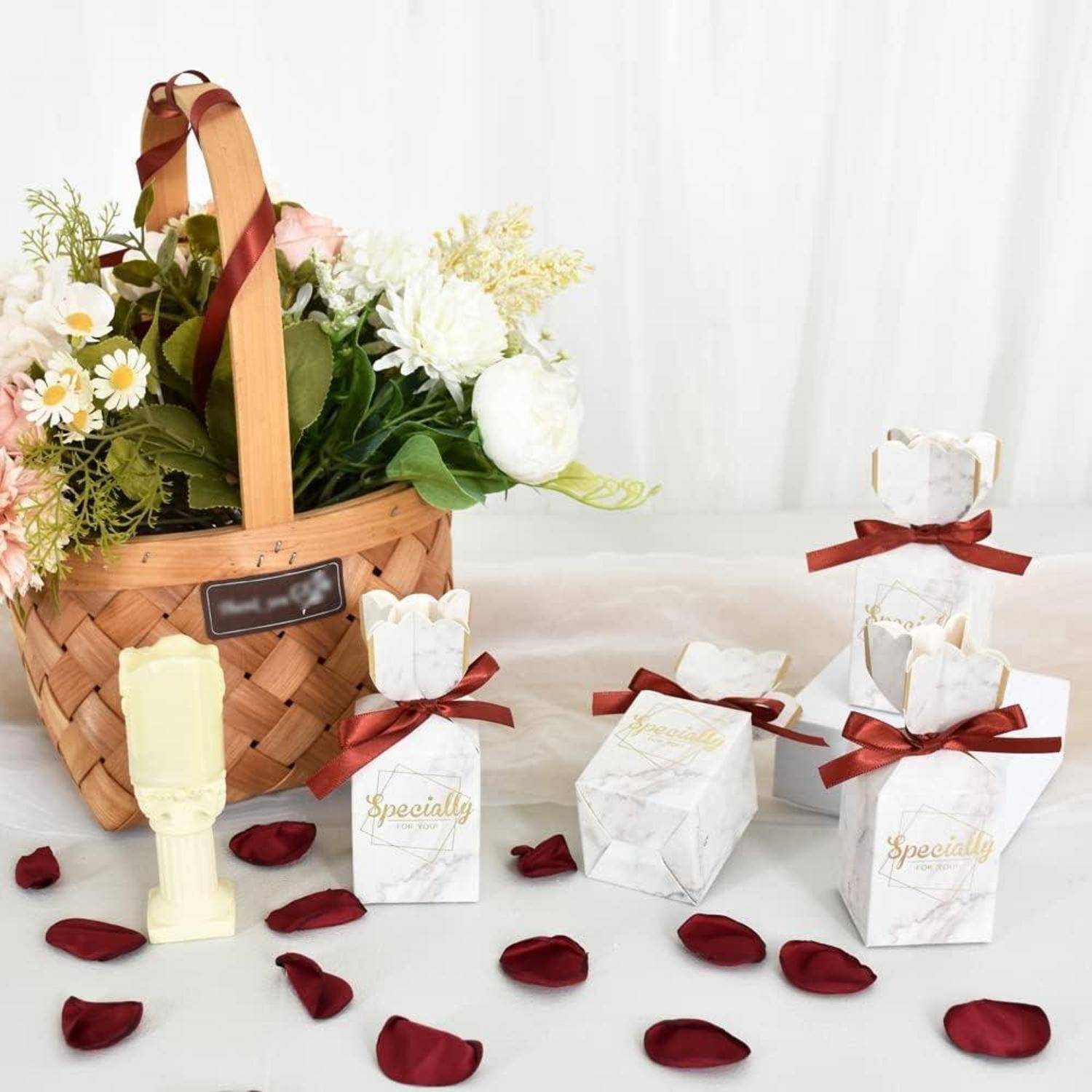 Ein Flechtkorb mit weißen und rosa Blumen neben vier weißen Geschenkboxen, die mit burgunderroten Bändern gebunden sind, auf einer Oberfläche, die mit Rosenblüten bestreut ist.