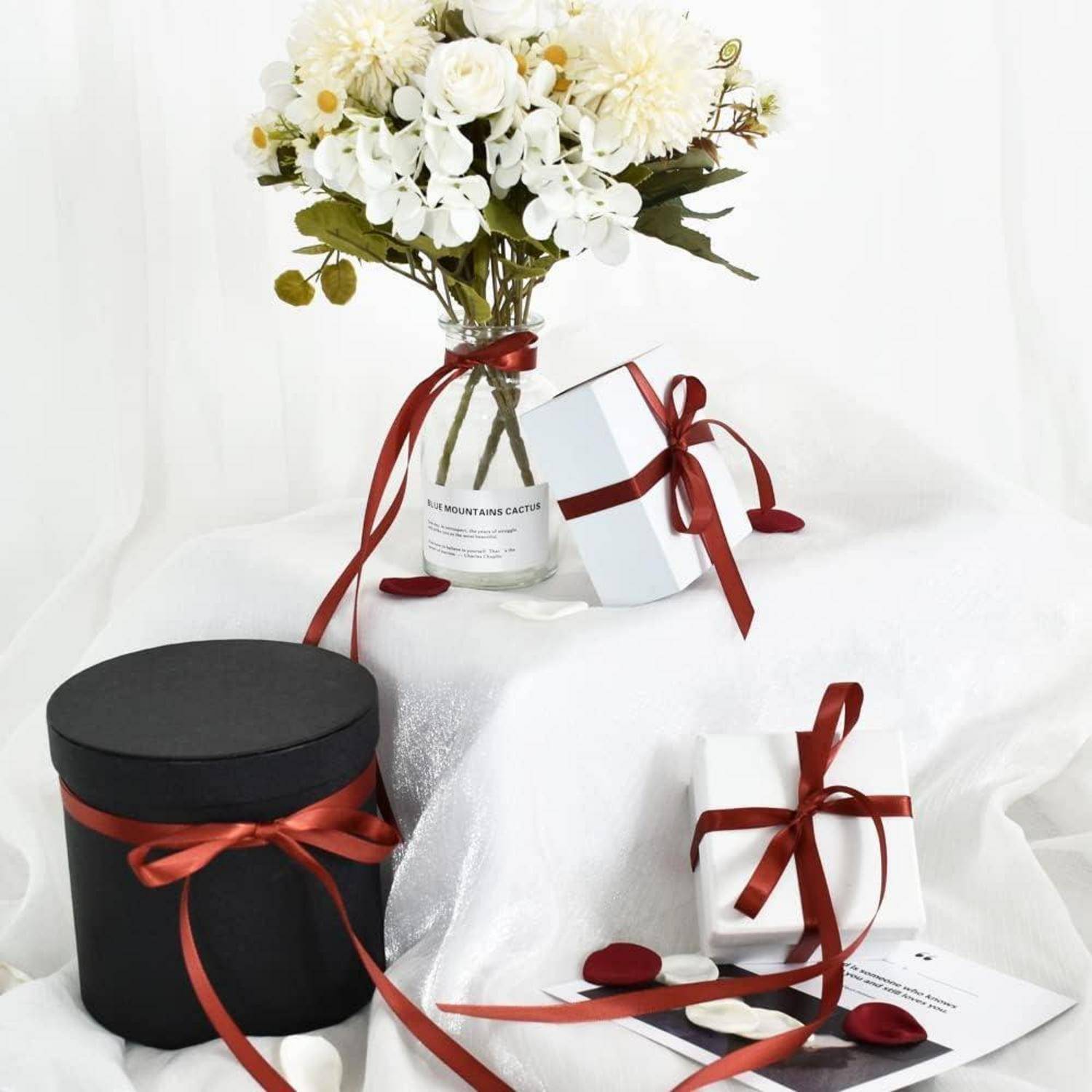 Eine schwarze runde Schachtel und eine weiße Schachtel, die mit roten Bändern umwickelt ist, neben einer Vase mit weißen Blumen, aufgestellt auf einem weißen Stoff.