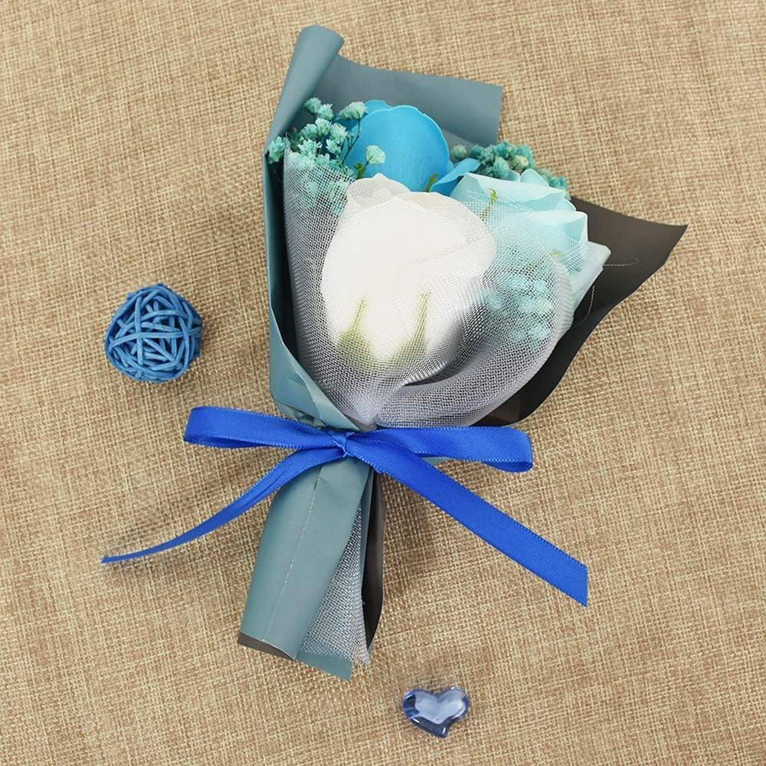 Ein kleiner Blumenstrauß mit weißen und blauen Rosen, in blaues Papier gewickelt und mit einer blauen Schleife gebunden, auf einer Juteoberfläche, neben einem blauen geflochtenen Ball und einem Herz.
