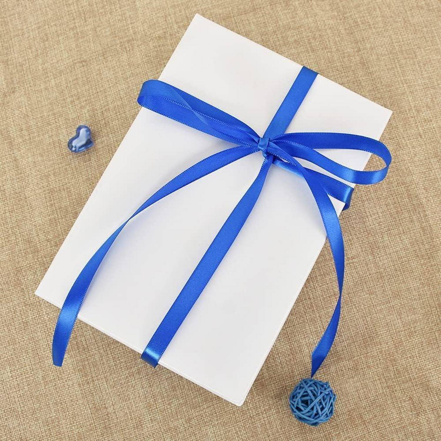 Eine weiße Geschenkbox mit einem blauen Band liegt auf einer beigen Oberfläche. In der Nähe befinden sich kleine blaue Dekorationsgegenstände: ein Herz und eine geflochtene Kugel.