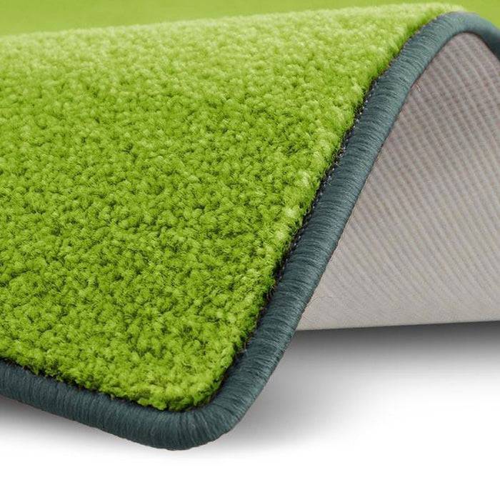 Eine Nahaufnahme einer hellgrünen Teppichecke, die leicht aufgerollt ist und dabei die weiße Unterseite und eine dunkelgrüne Einfassungskante zeigt.