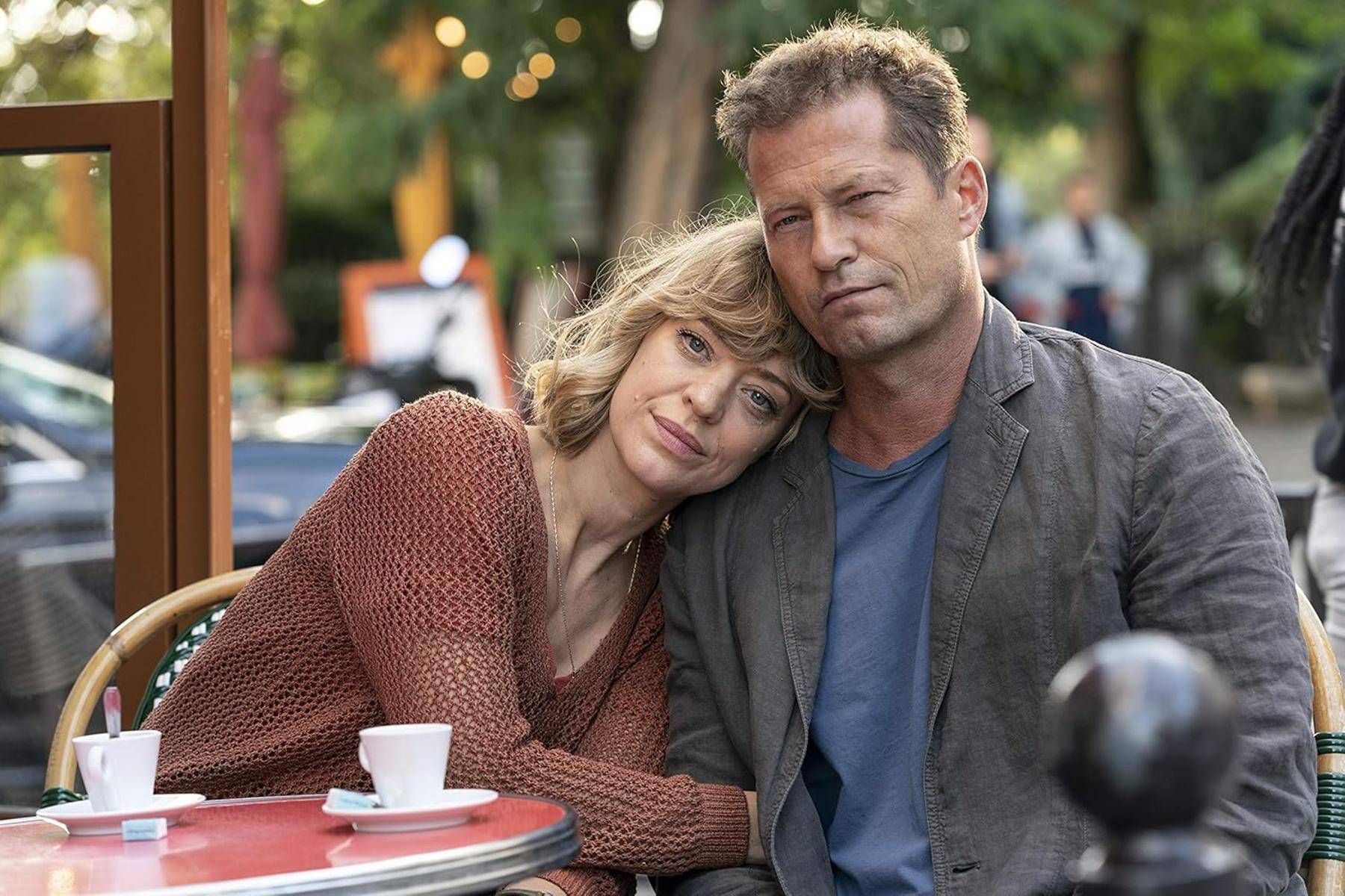 Ein Paar sitzt im Freien, wobei die Frau ihren Kopf auf der Schulter des Mannes ruhen lässt. Eine Tasse steht auf dem Tisch, und im Hintergrund verschwommenes Grün zu sehen ist.