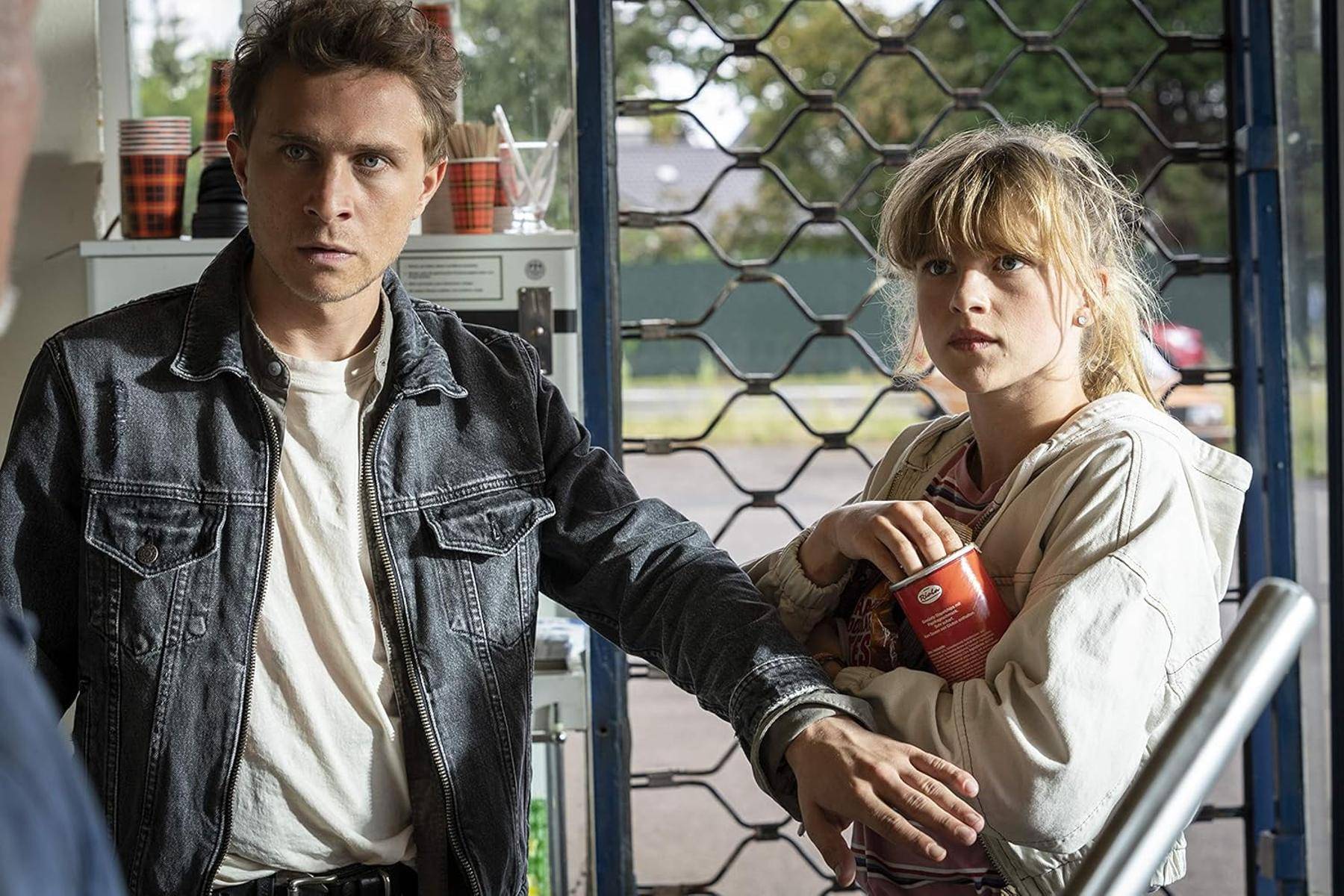 Ein Mann und eine Frau stehen dicht beieinander in einem Raum und blicken aufmerksam auf etwas draußen. Die Frau hält ein Getränk, und der Mann trägt eine Jeansjacke.