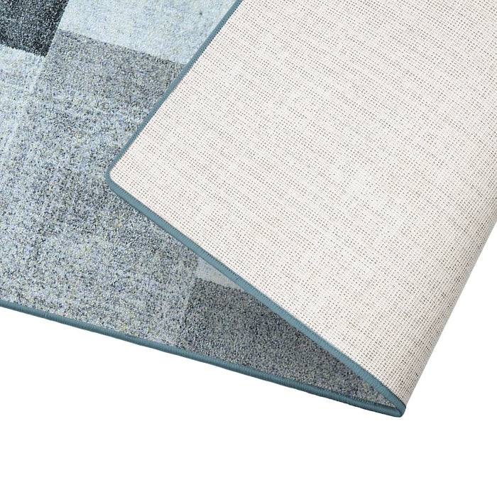 Ein rechteckiger Teppich mit einem geometrischen Muster in verschiedenen Grautönen ist an einer Ecke umgeschlagen und zeigt dabei seine beige Unterseite.