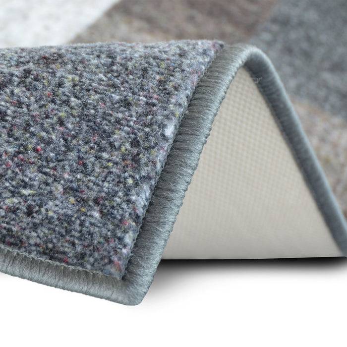 Ein Nahaufnahmebild einer Ecke eines grauen, strukturierten Teppichs, der umgefaltet ist und die rutschfeste Rückseite auf einer glatten Oberfläche zeigt.