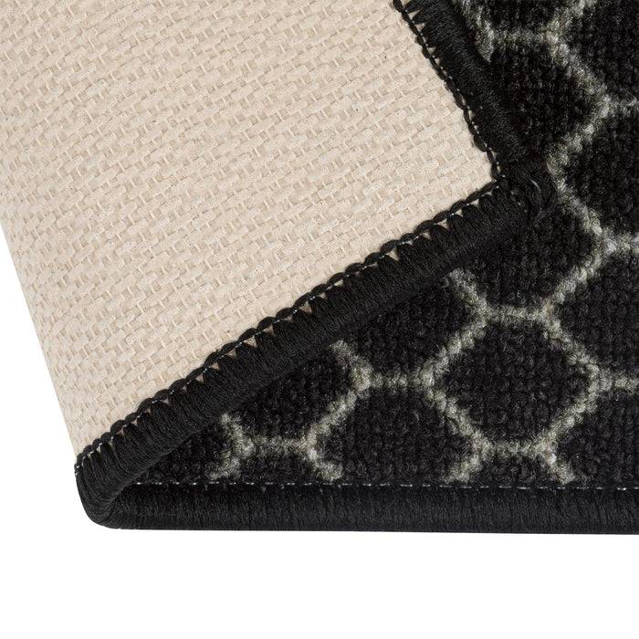 Eine Nahaufnahme einer Ecke eines schwarz-beigen Flachteppichs, die geometrische Muster auf der Vorderseite und eine strukturierte Unterseite für Rutschfestigkeit zeigt.
