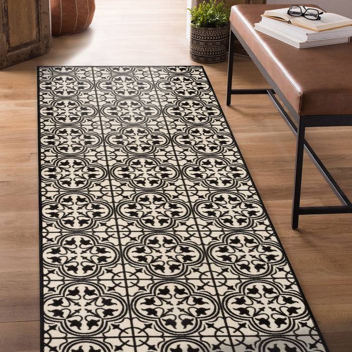 Ein schwarz-weißer gemusterter Teppich erstreckt sich entlang eines Holzbodens neben einer braunen Bank mit Büchern. Im Hintergrund steht eine Topfpflanze.