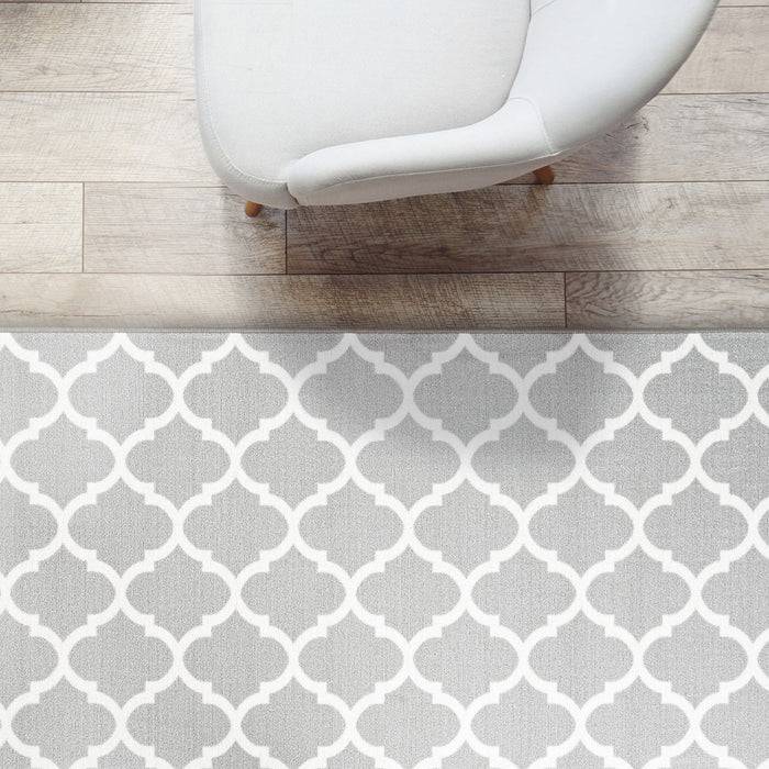 Ein gebogener weißer Stuhl steht auf Holzdielen neben einem grauen Teppich mit weißem geometrischem Muster.