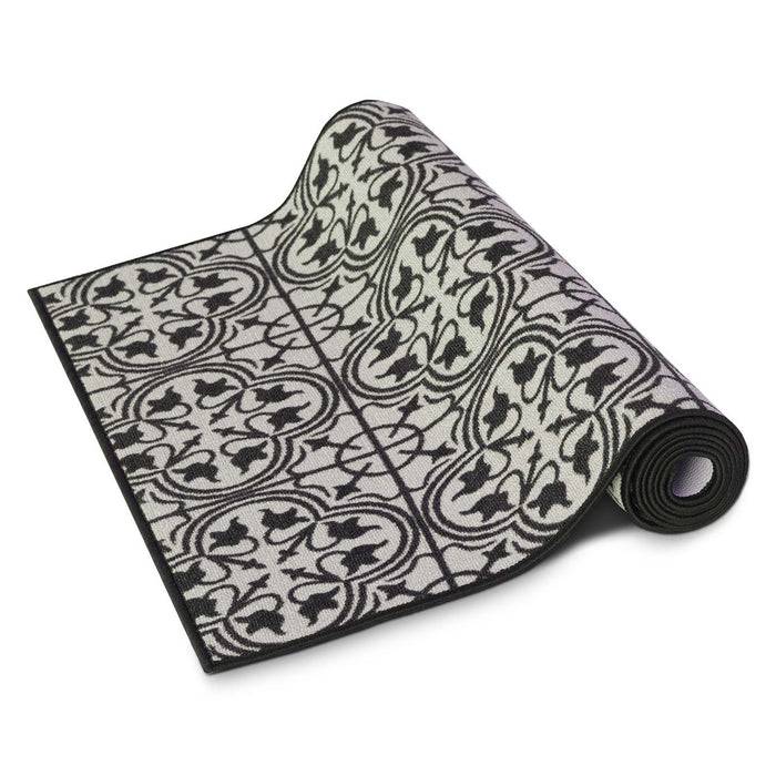 Ein aufgerollter dekorativer schwarz-weißer Teppich mit einem kunstvollen Blatt- und Rankenornament. Der Teppich ist teilweise entrollt und liegt auf einer einfarbigen Oberfläche.