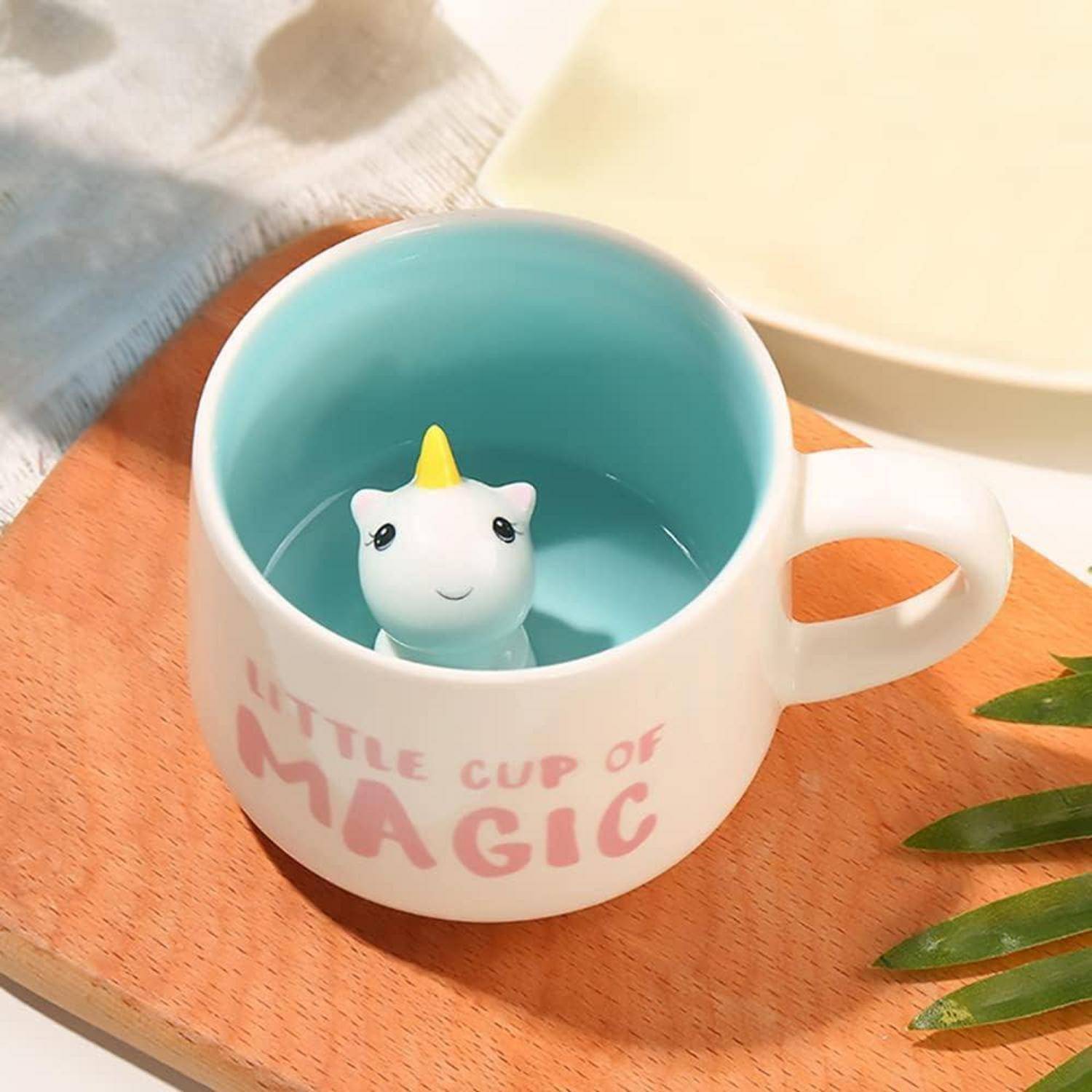 Eine weiße Tasse mit der Aufschrift 'Little Cup of Magic', in der eine kleine Keramik-Einhorn-Figur sitzt, platziert auf einer Holzoberfläche mit weichem Licht.