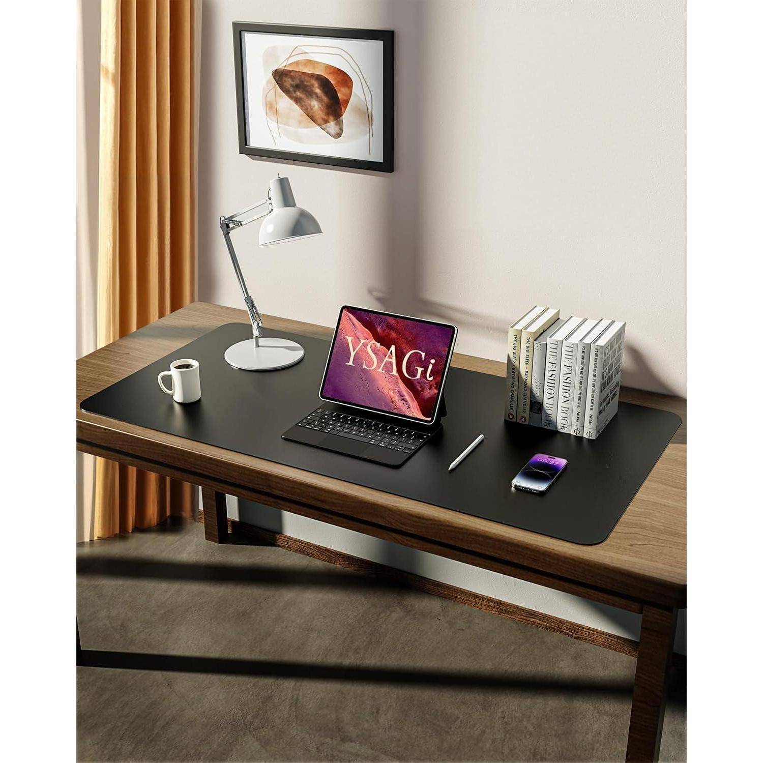 Ein ordentlich aufgeräumter Schreibtisch mit einem Laptop, auf dessen Bildschirm ‚YSAGi' angezeigt wird, einer Lampe, einem Smartphone, einem Stift und einem Stapel Büchern, mit gerahmter Kunst an der Wand.
