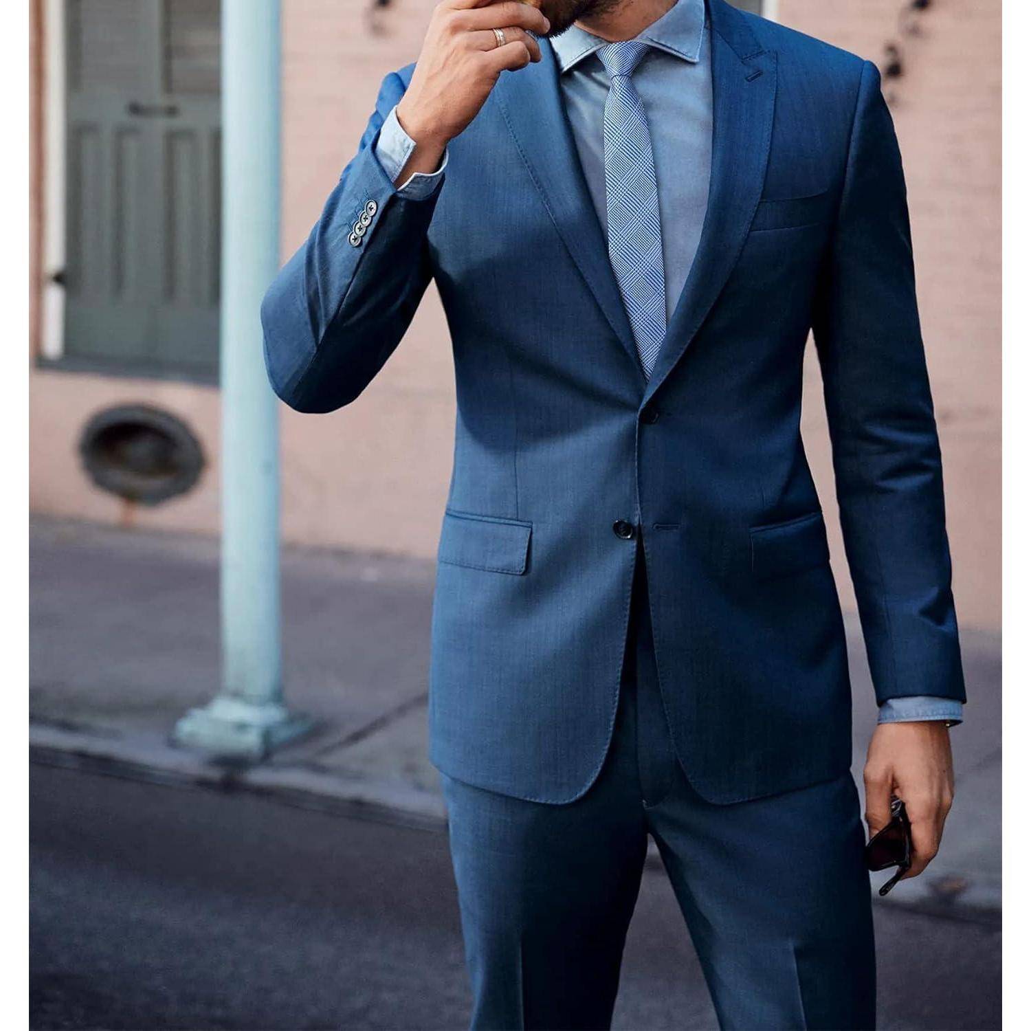 Ein Mann in einem blauen Anzug mit einem hellblauen Hemd und einer Krawatte steht im Freien in der Nähe eines Gebäudes. Er hält ein Smartphone in seiner rechten Hand.
