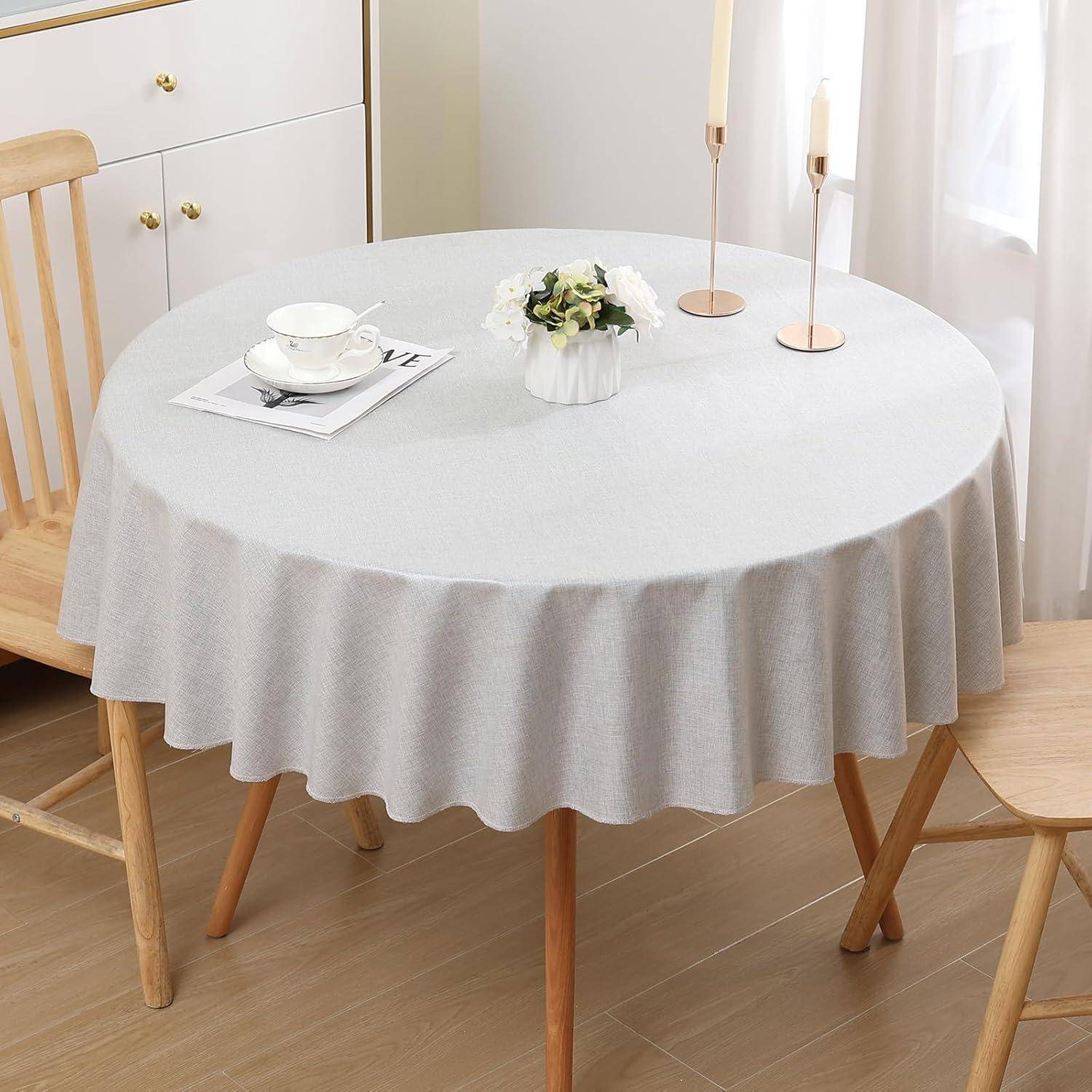 Ein runder Tisch mit einer hellgrauen Tischdecke ist mit einer weißen Teetasse, Untertasse und einer kleinen weißen Vase mit Grünpflanzen in einem hellen Raum gedeckt.