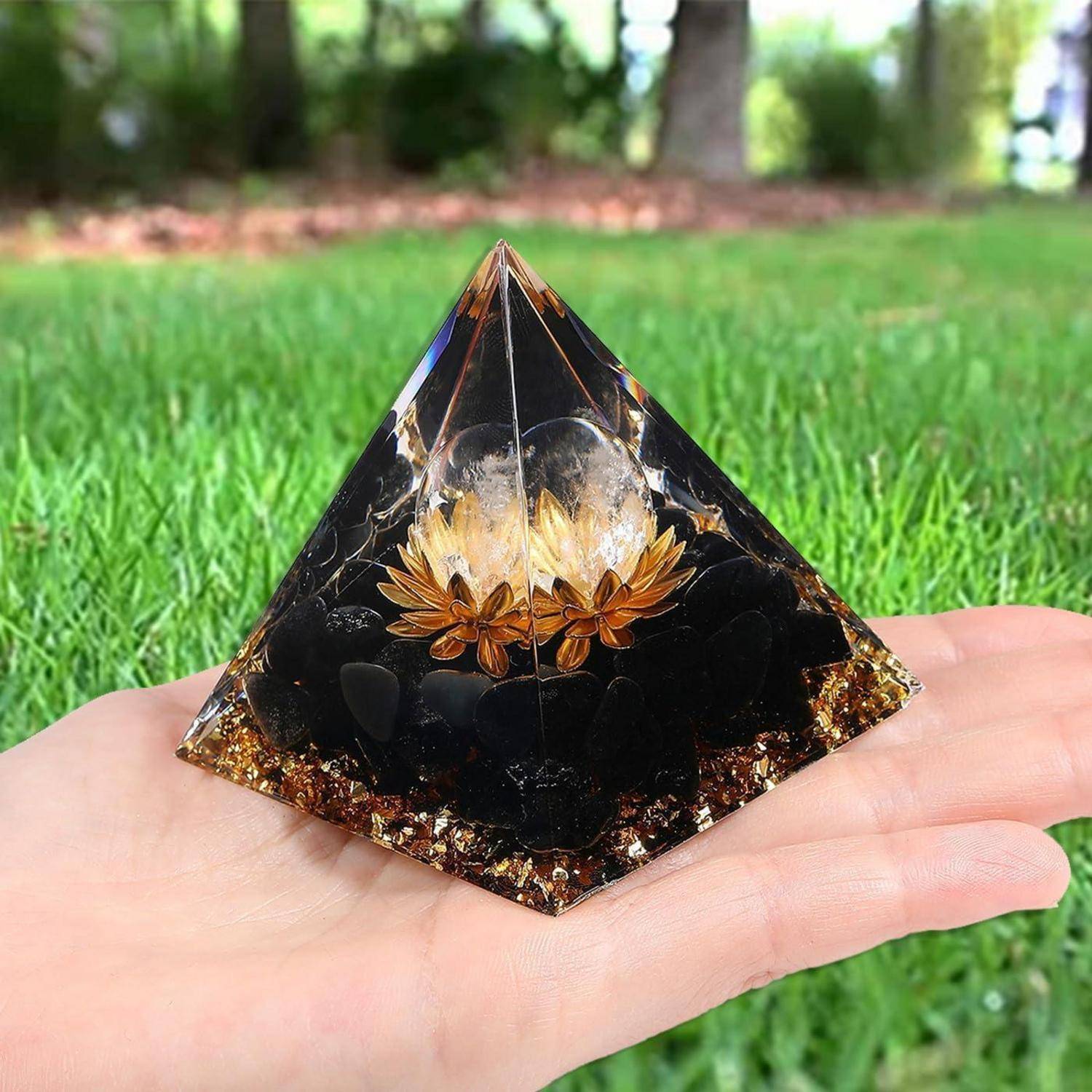 Eine Hand hält eine dekorative Pyramide mit bernsteinfarbenen und schwarzen Schichten, in der sich ein Blumendesign befindet. Der Hintergrund zeigt eine Rasenfläche und Bäume.