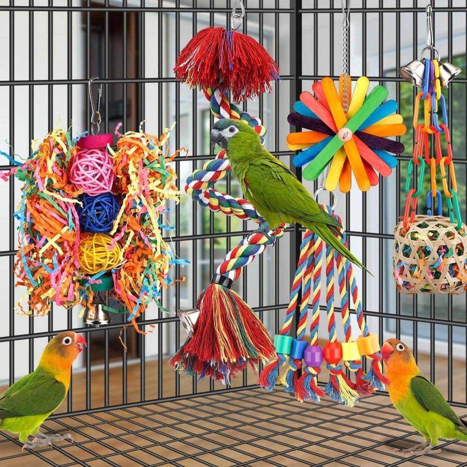 Ein grüner Papagei in einem Käfig mit bunten hängenden Spielzeugen und Seilen. Zwei kleinere Vögel befinden sich unten außerhalb des Käfigs.
