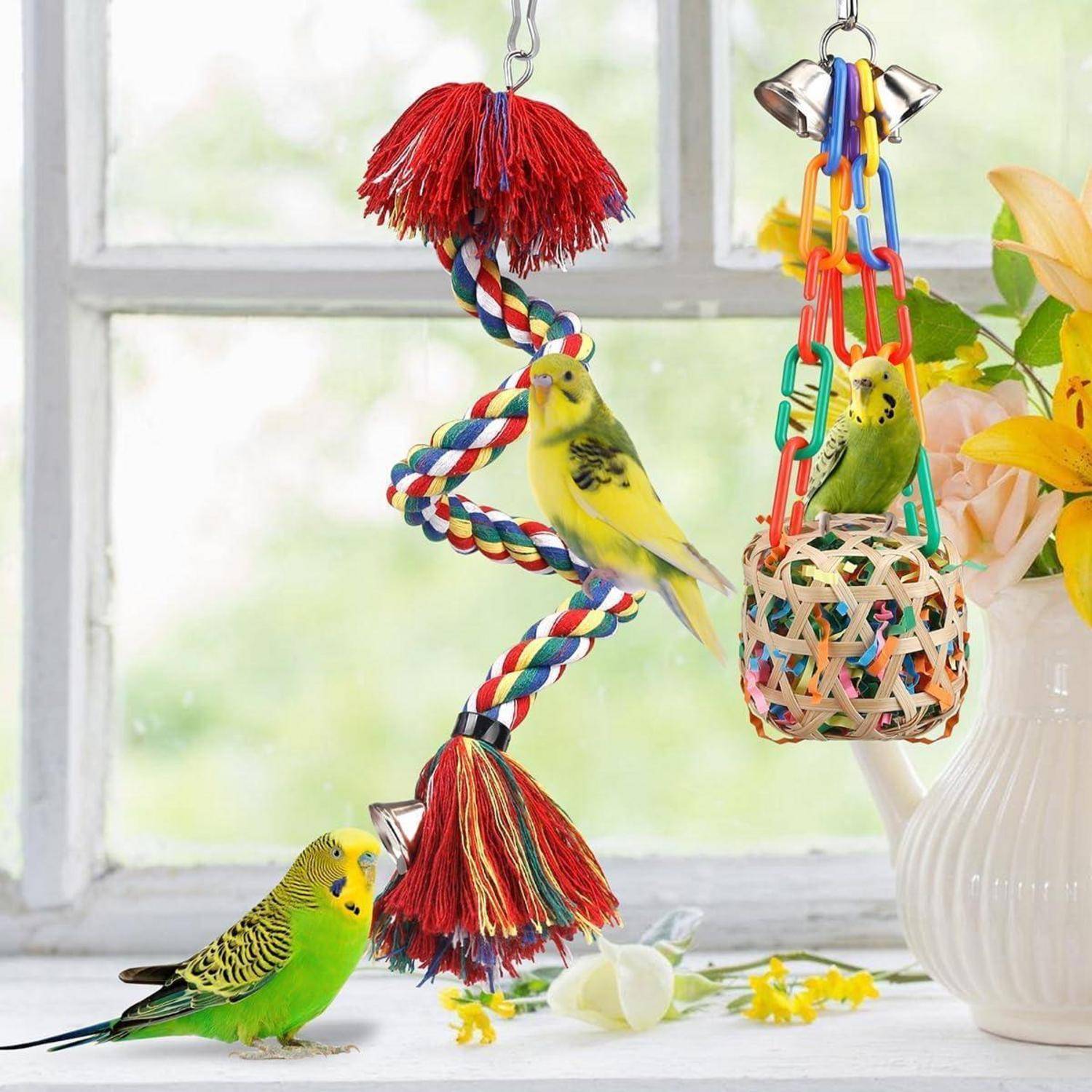 Ein gelber und ein grüner Wellensittich spielen mit bunten hängenden Spielzeugen neben einem Fenster. Gelbe Blumen sind rechts sichtbar.