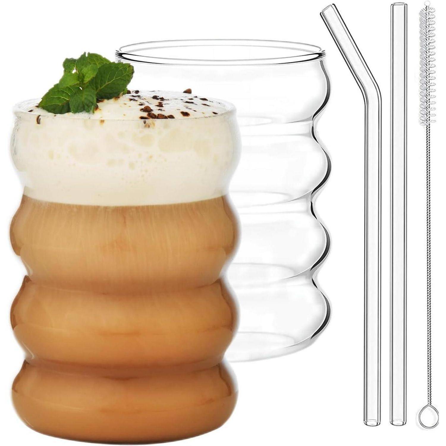 Eine gewellte Glasstasse gefüllt mit Eiskaffee, gekrönt von Schaum und Minze. Daneben stehen ein leeres Trinkglas, Glastrinkhalme und eine Reinigungsbürste.