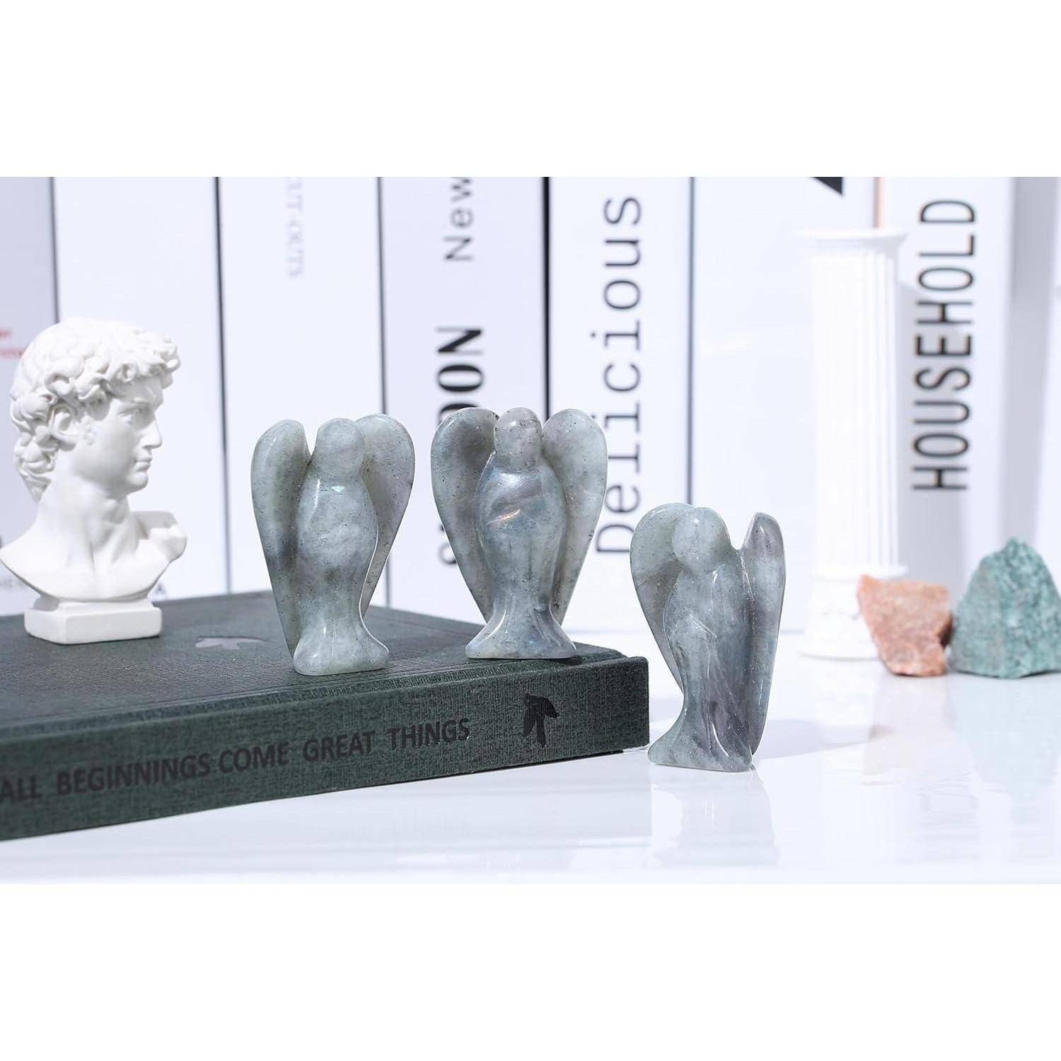 Drei geschnitzte Steinengel-Statuen sind auf einem Bücherregal mit verschiedenen Büchern, einer kleinen Büste und Kristallen ausgestellt, was eine elegante Atmosphäre schafft.