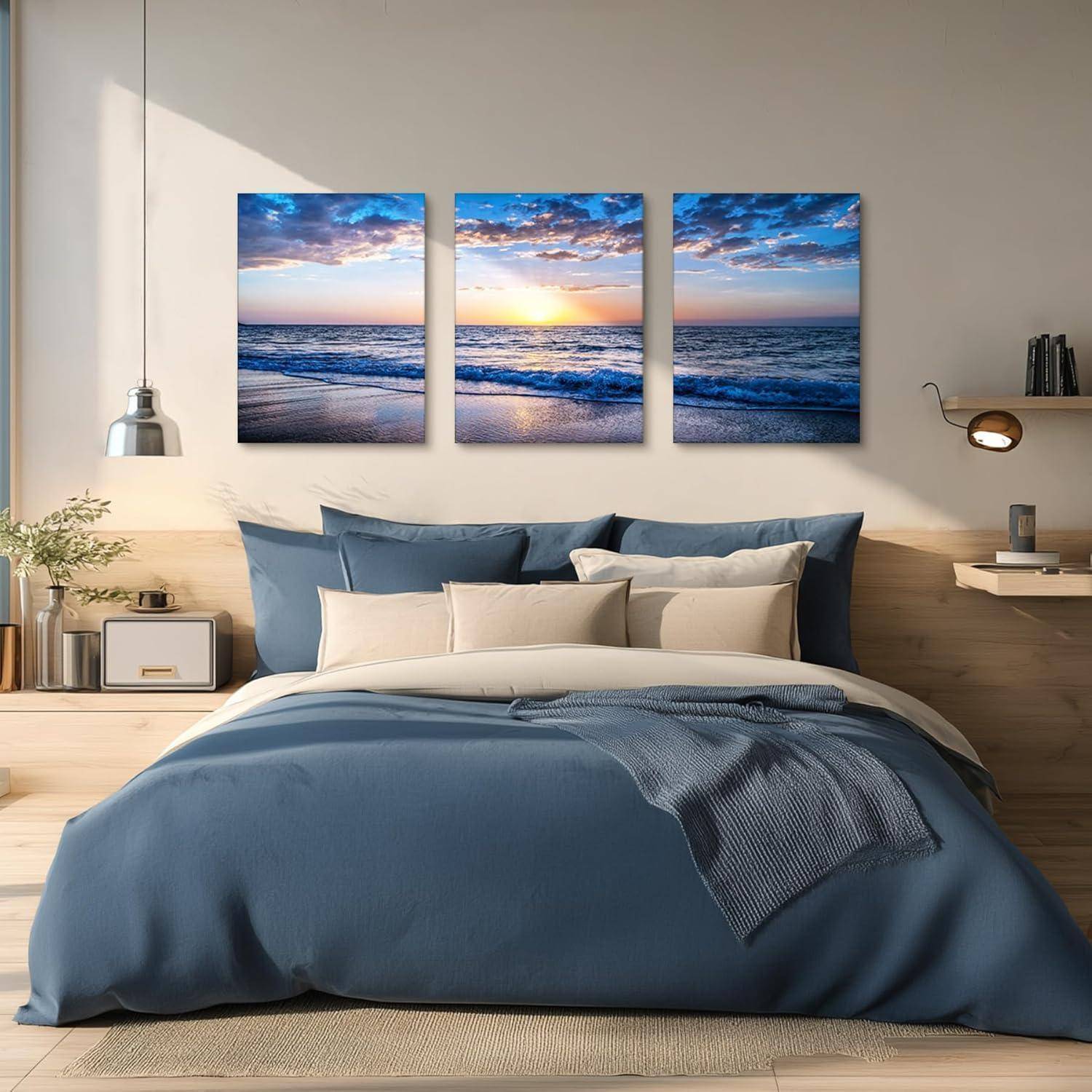Ein gemütliches Schlafzimmer mit einem blauen Bett, beigen Kissen, Holzregalen und einem dreiteiligen Sonnenuntergangs-Meereskunstwerk an der Wand über dem Bett.