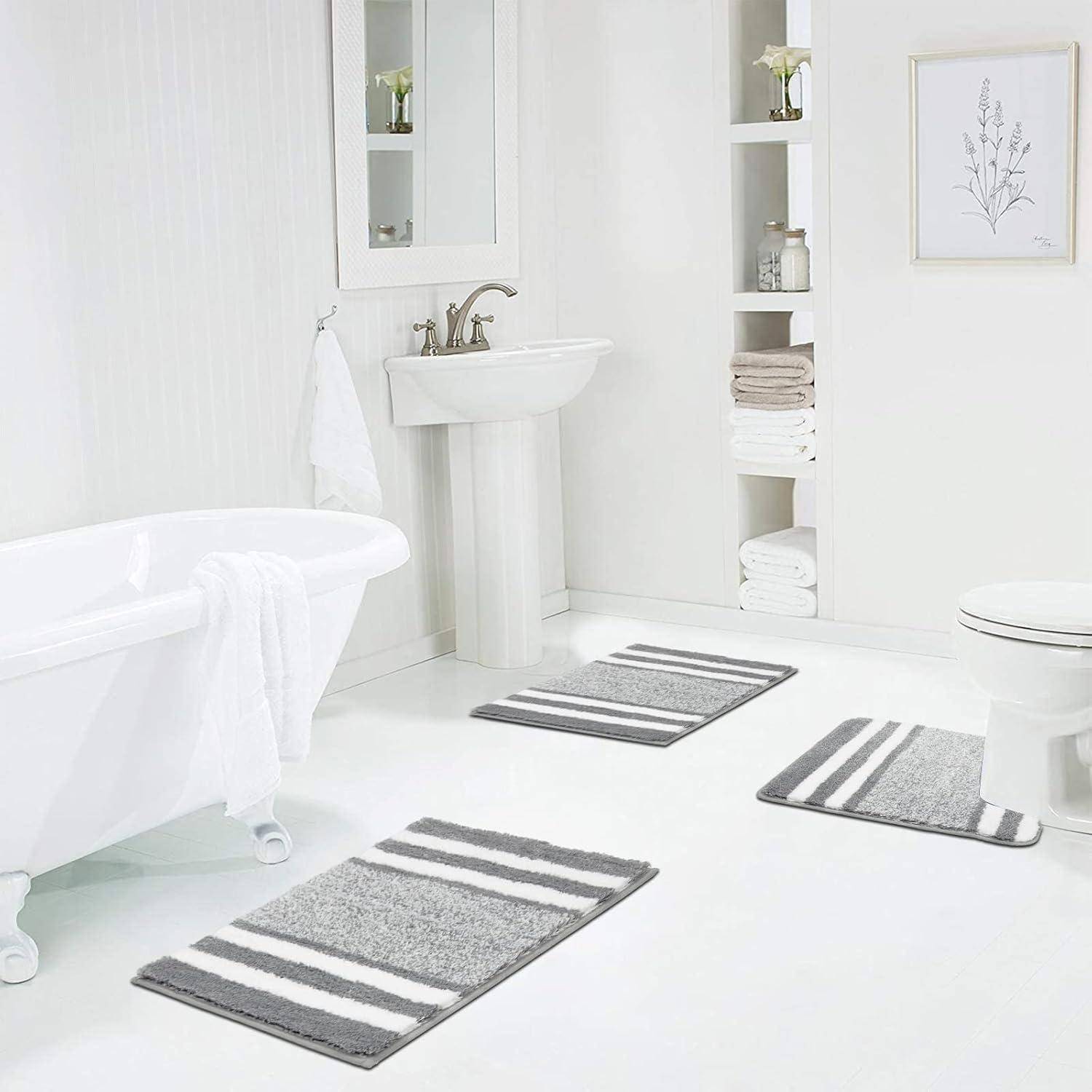 Ein weißes Badezimmer mit einer Badewanne, einem Standbecken und einer Toilette. Regale tragen dekorative Gegenstände. Drei gestreifte graue Badematte auf dem Boden.