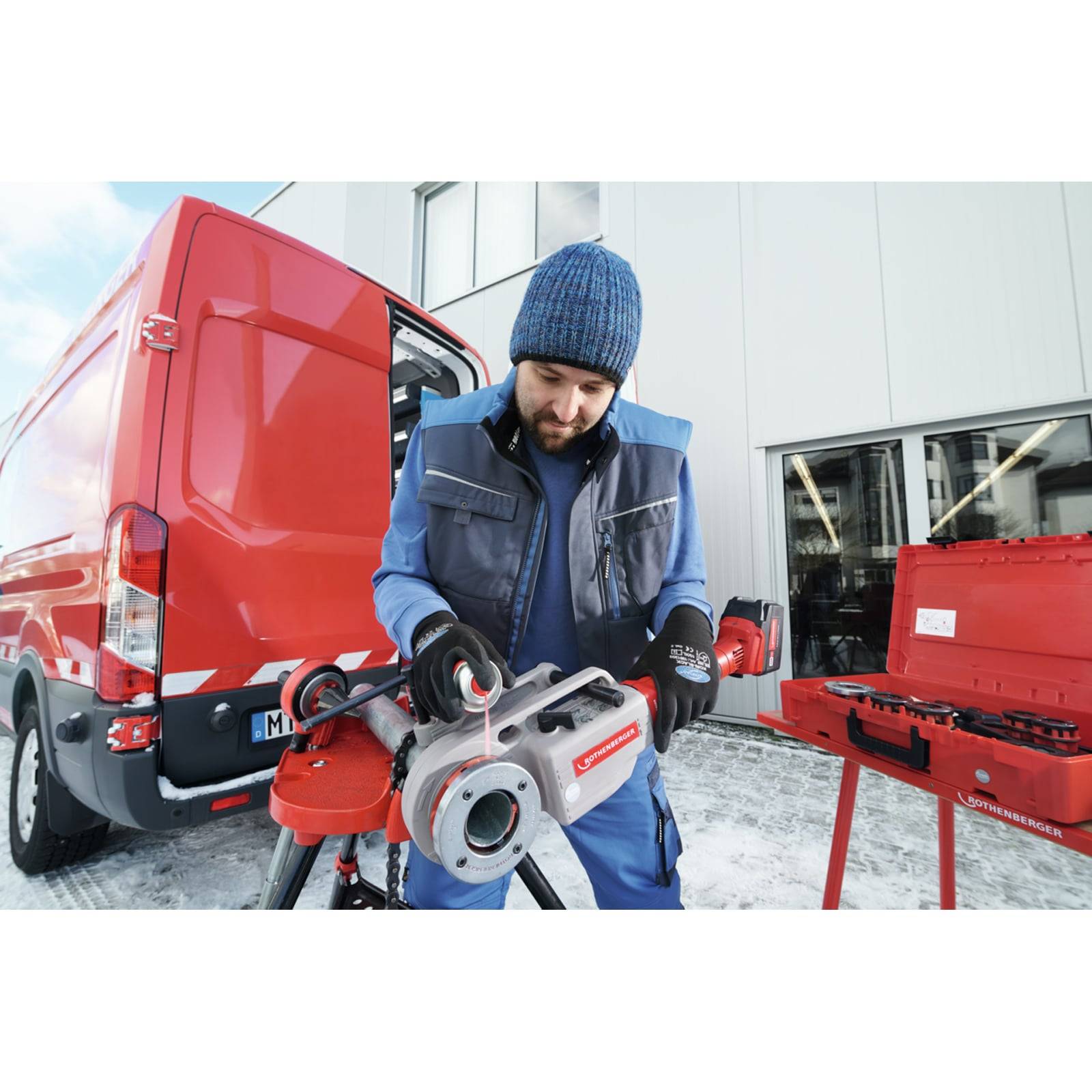 Eine Person in Winterkleidung bedient eine Rohgewindemaschine neben einem roten Van und einem Werkzeugkoffer, vor einem Industriegebäude.