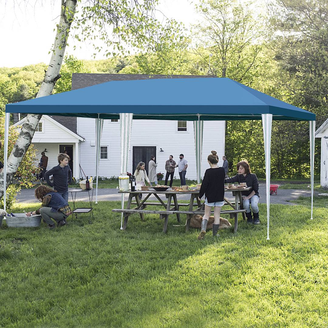 Menschen versammeln sich unter einem blauen Baldachin in einem grasbedeckten Garten, einige sitzen an einem Picknicktisch, andere stehen und unterhalten sich, mit Bäumen im Hintergrund.