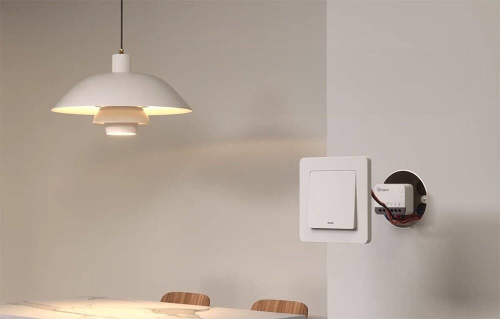 Ein moderner Essbereich zeichnet sich durch eine hängende weiße Lampe über einem Tisch mit Holzstühlen aus. Ein quadratisches weißes Gerät ist an der nahegelegenen Wand montiert.