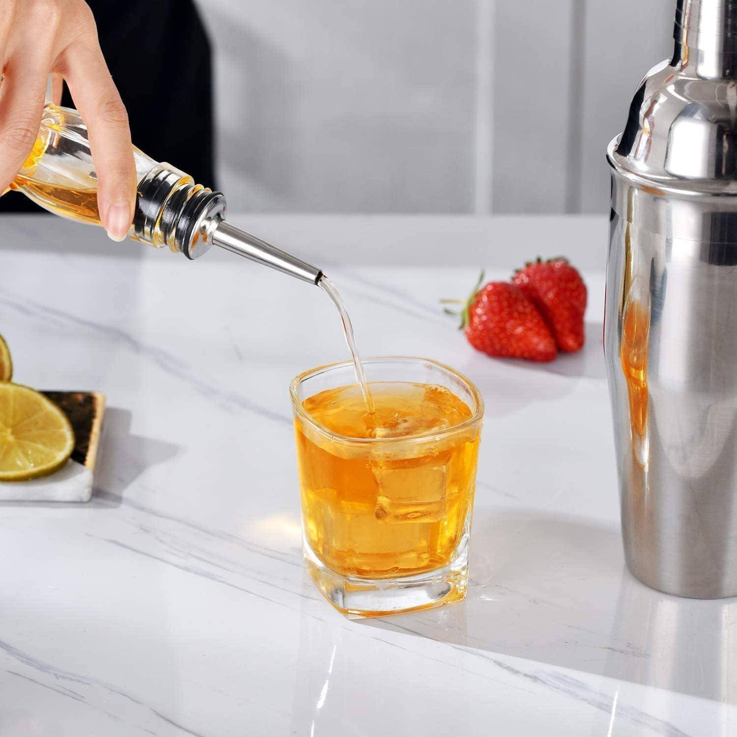 Eine Hand gießt eine goldene Flüssigkeit in ein Glas auf einer Marmorarbeitsplatte. In der Nähe liegen Zitronenscheiben und Erdbeeren, rechts daneben steht ein glänzender Shaker.