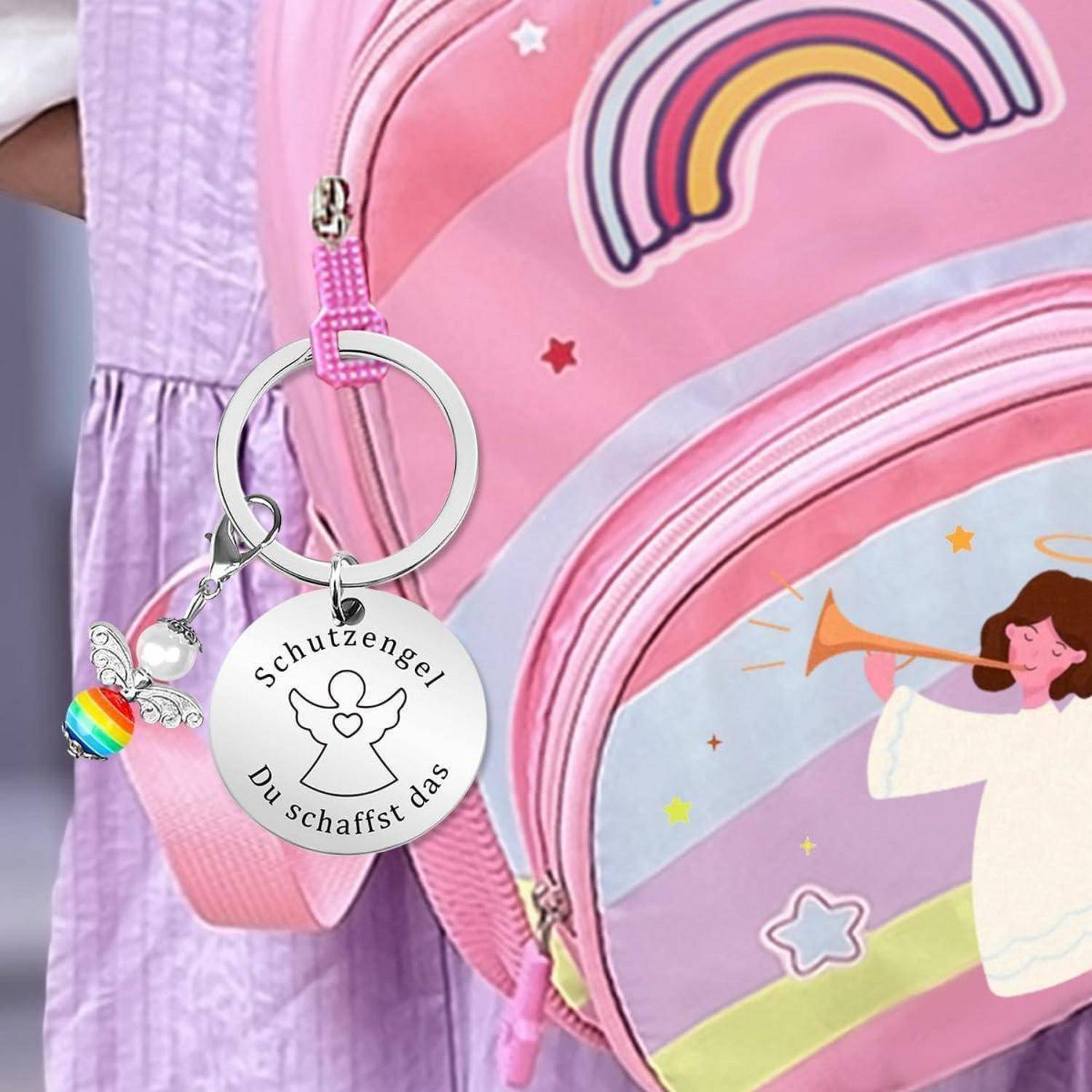 Ein rosa Rucksack mit Regenbogen- und Engelsgrafiken, der einen Schlüsselanhänger trägt mit der Aufschrift „Schutzengel Du schaffst das