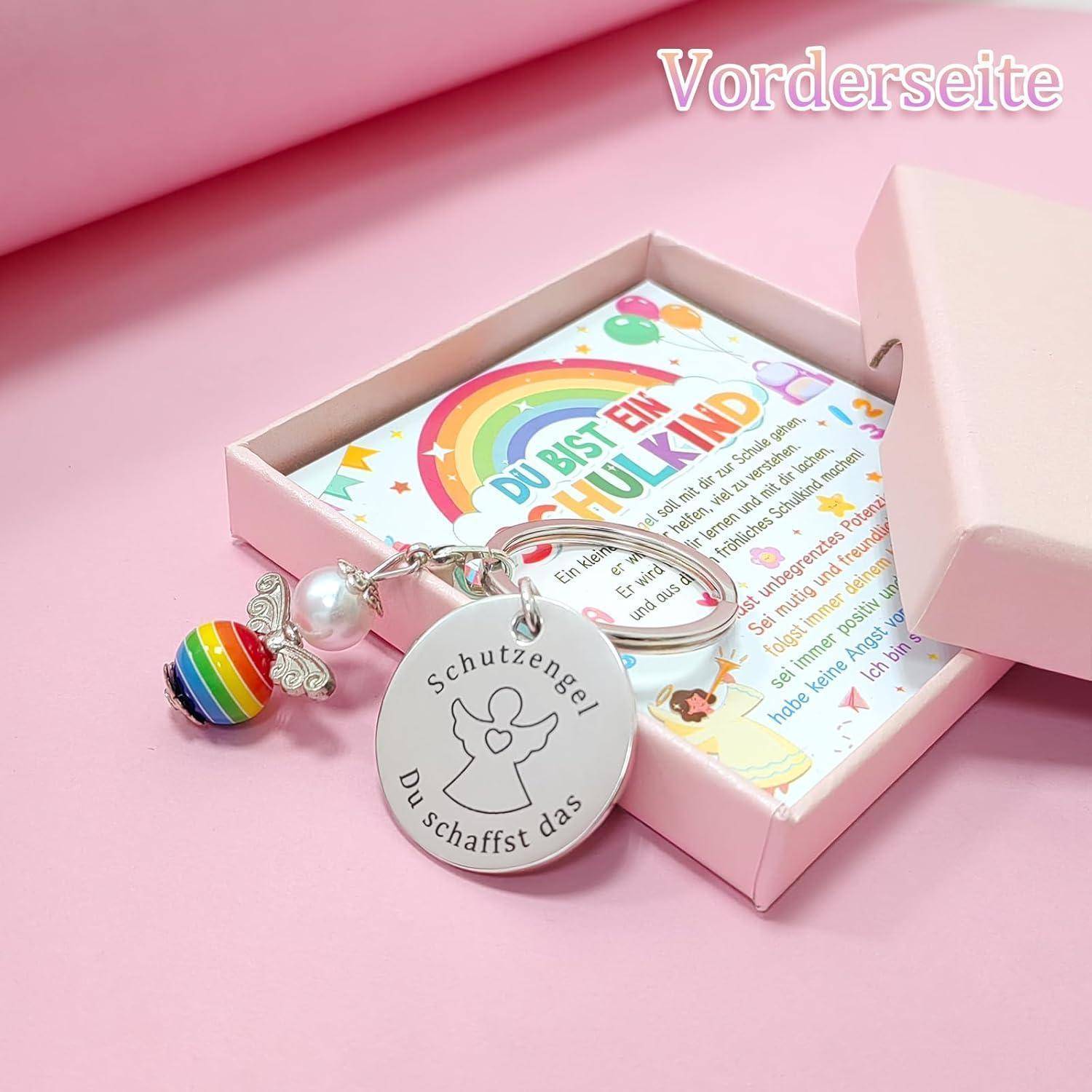 Rosa Geschenkbox mit einem 'Schutzengel Du schaffst das' Schlüsselanhänger und einem bunten Herz, geöffnet mit einer Karte, auf der ein Regenbogen und der Satz 'Du bist ein Schulkind' zu sehen ist.