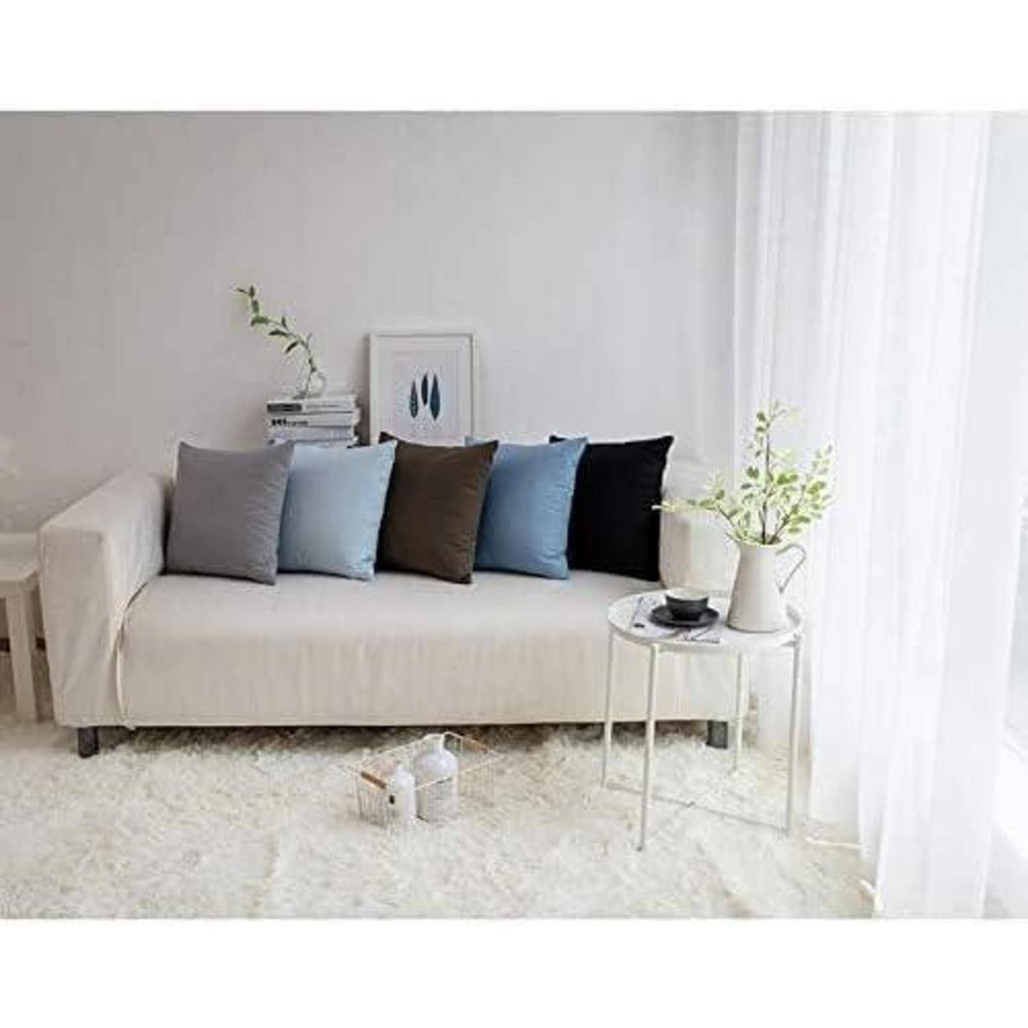 Ein weißes Sofa mit fünf Kissen in Blau- und Grautönen steht an einer weißen Wand. Daneben befindet sich ein kleiner runder Tisch mit einer Pflanze und einer Tasse.