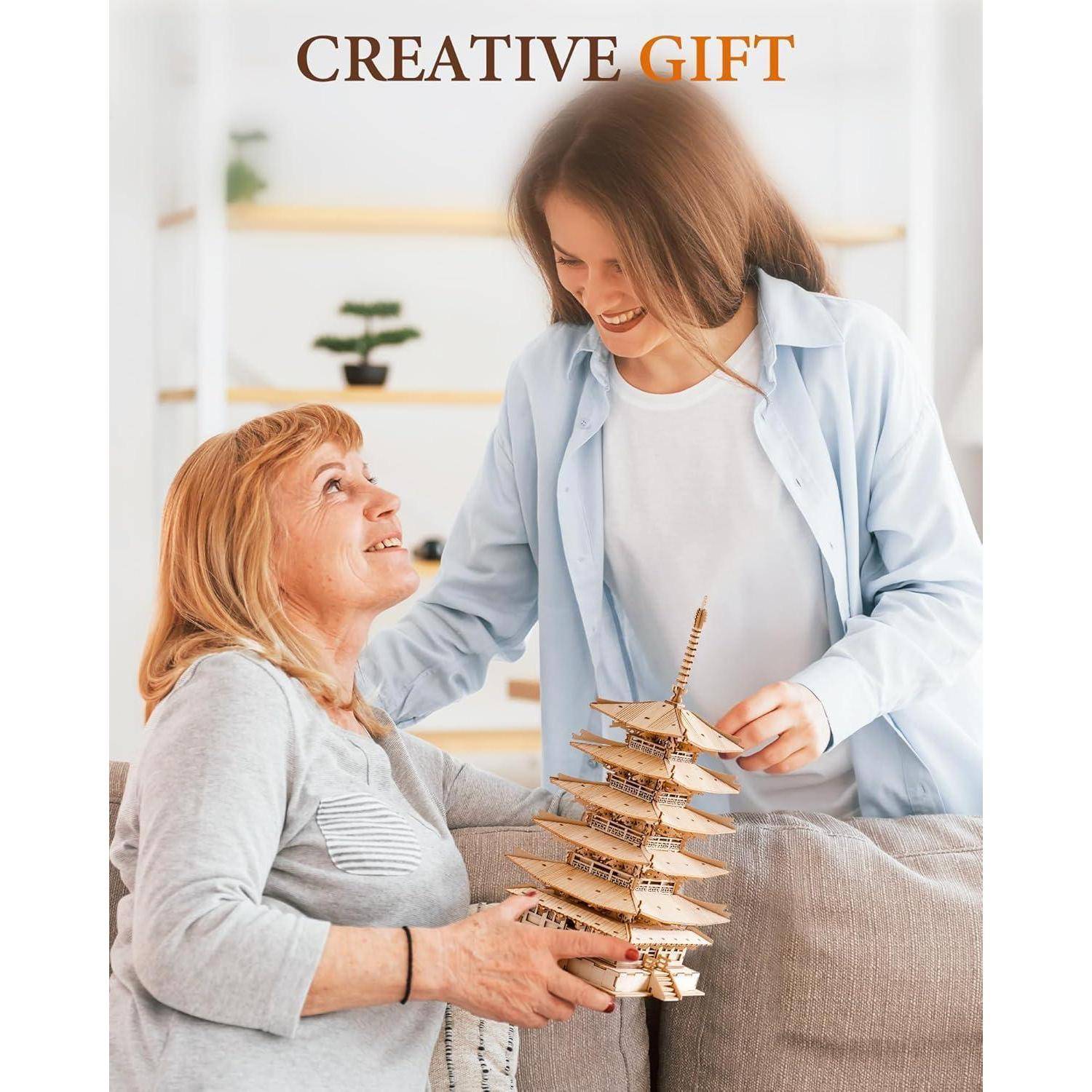 Eine Frau auf einem Sofa lächelt und hält ein Holzpagoden-Modell, während eine jüngere Frau daneben steht und es ihr überreicht. Text lautet: ‚Kreatives Geschenk'.