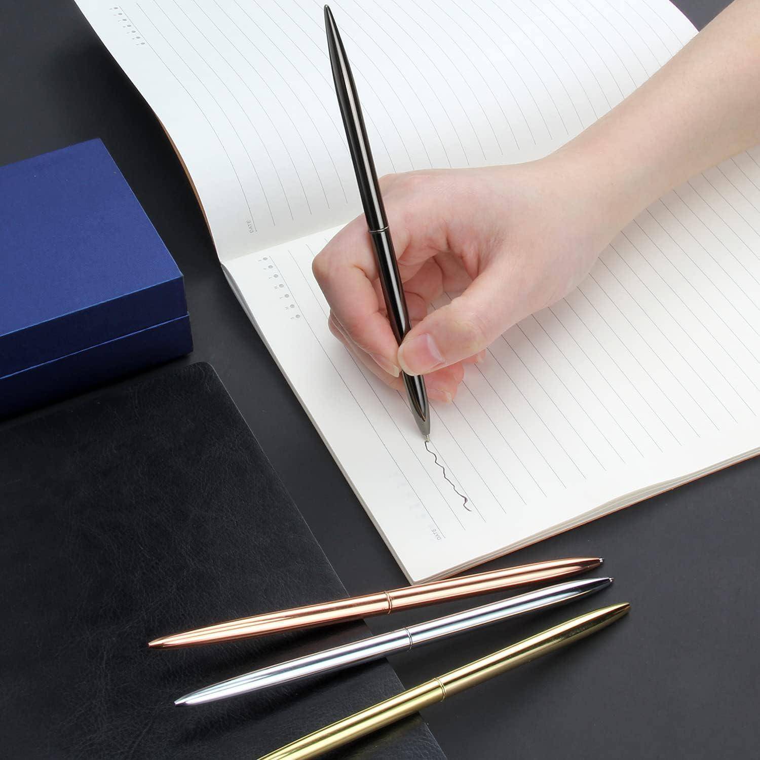 Eine Hand, die einen schwarzen Stift über ein geöffnetes liniertes Notizbuch hält und kurz davor ist zu schreiben, mit drei Stiften und zwei geschlossenen Notizbüchern daneben auf einem Schreibtisch.