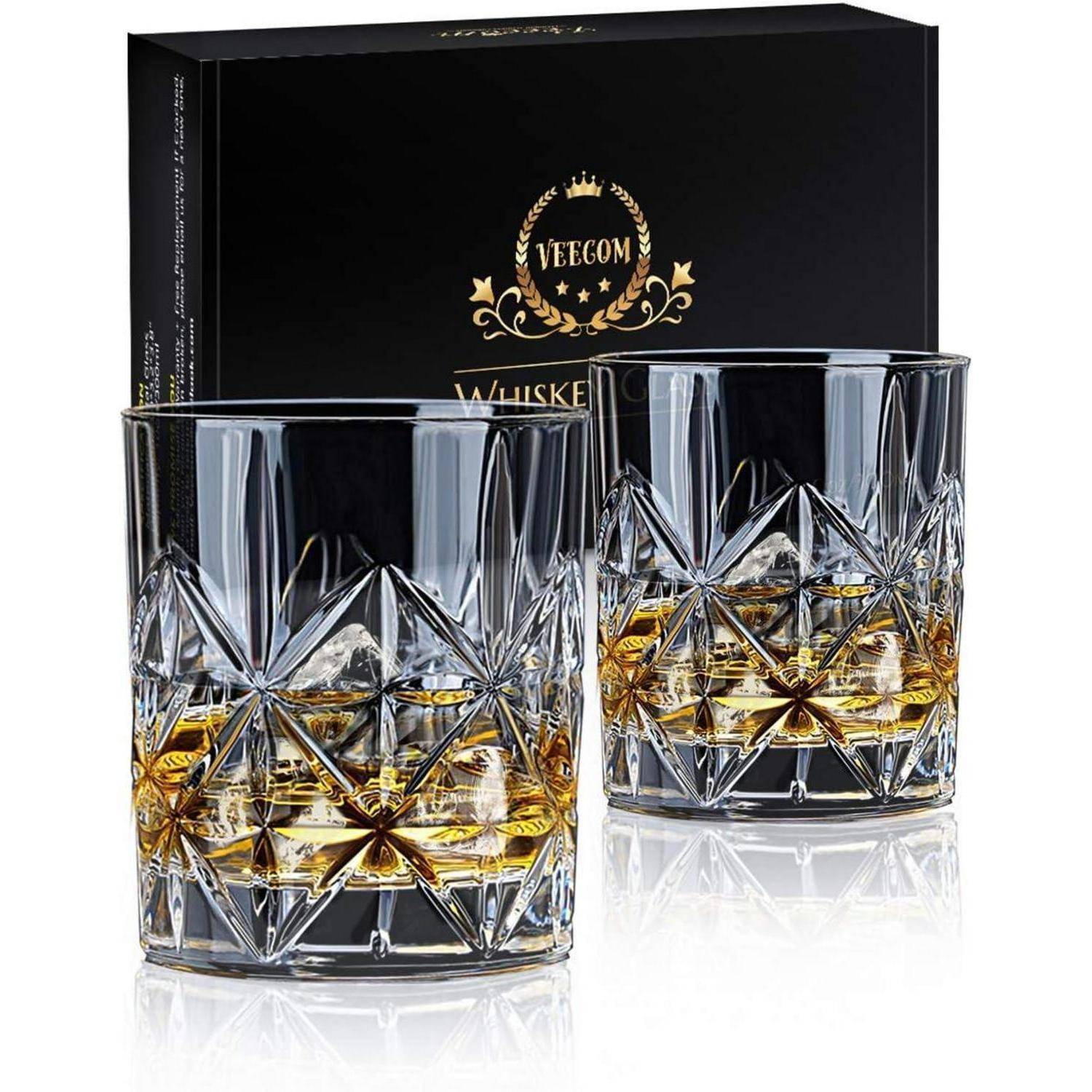 Zwei Kristall-Whiskygläser mit kunstvollen Mustern, zur Hälfte mit bernsteinfarbener Flüssigkeit gefüllt, vor einer schwarzen Geschenkbox mit goldenen Akzenten.