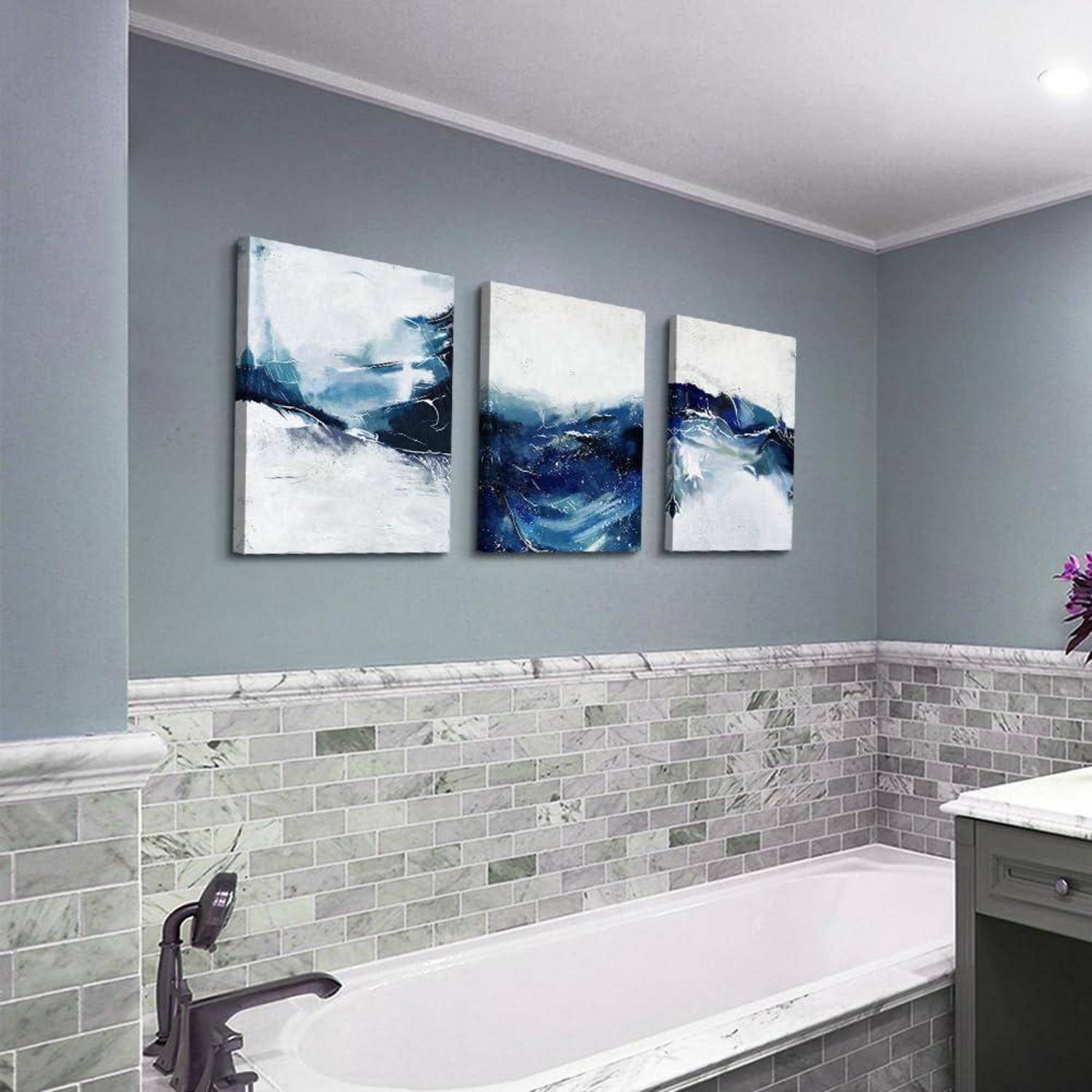 Ein modernes Badezimmer mit einer weißen Badewanne, grauen Fliesenwänden und drei abstrakten blau-weißen Gemälden über der Badewanne.
