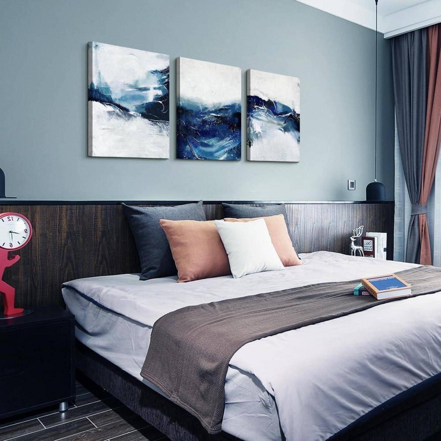 Modernes Schlafzimmer mit einem Bett, das graue und pfirsichfarbene Kissen, eine dunkle Decke und abstrakte Kunst über dem Kopfteil aufweist; Uhr und Lampe auf dem Nachttisch.