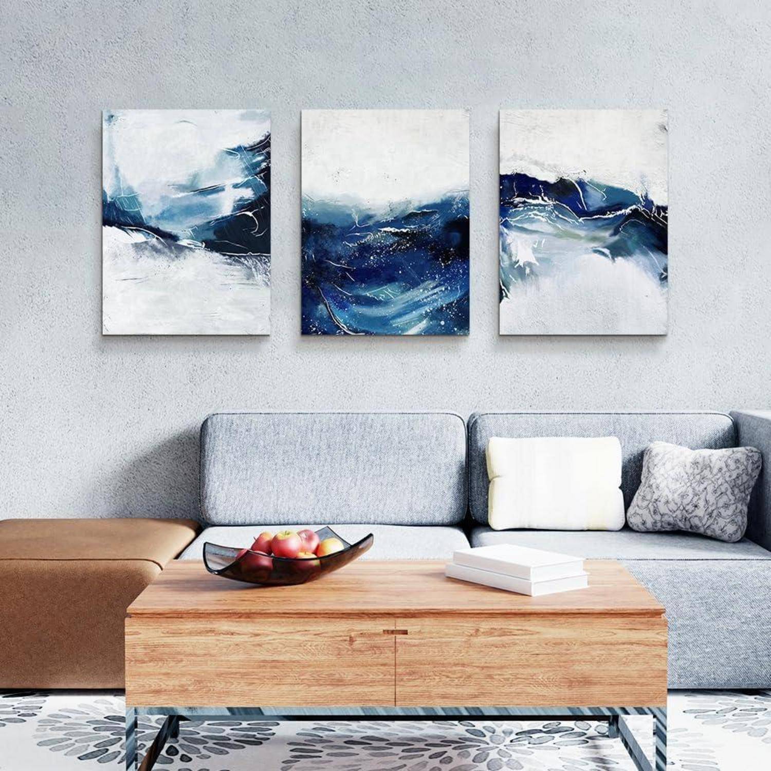 Ein moderner Wohnraum zeigt ein hellgraues Sofa, einen Couchtisch aus Holz mit einer Obstschale und drei abstrakte Gemälde in Blau und Weiß an der Wand.